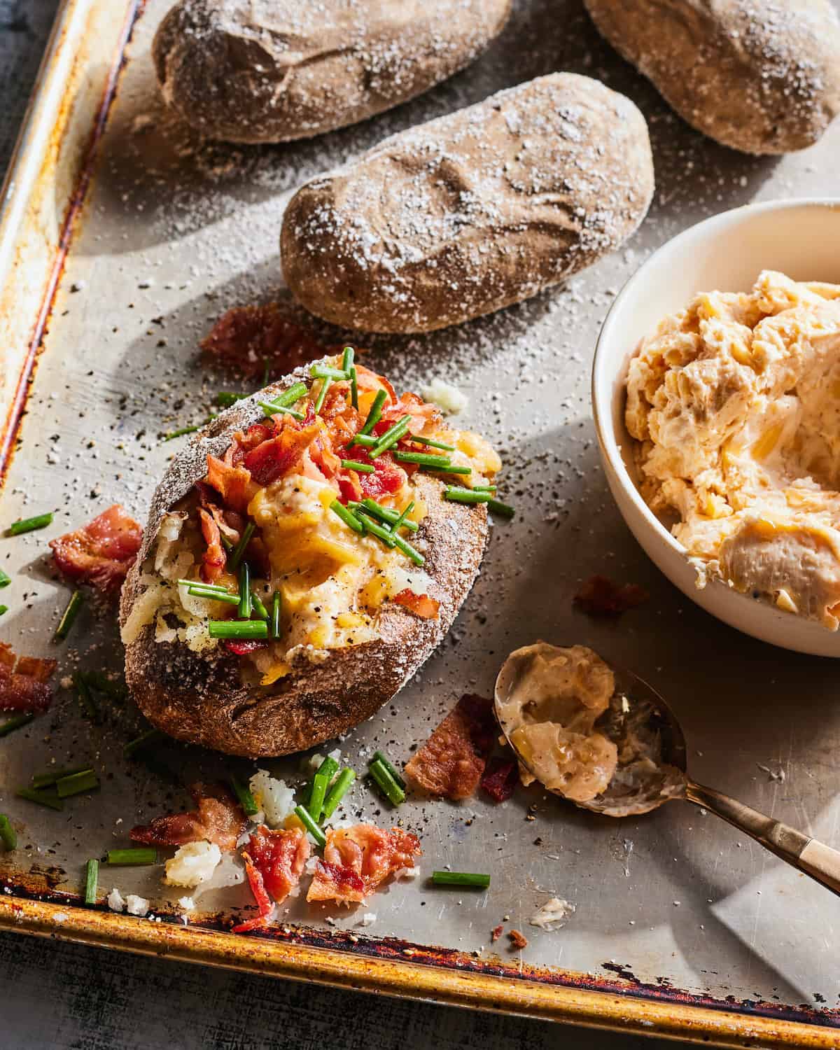 A baking sheet pan with baked potatoes rubbed with salt, one of them topped with garlic cheese butter, crispy bacon bits and chives, along with a bowl of garlic cheese butter on the side.