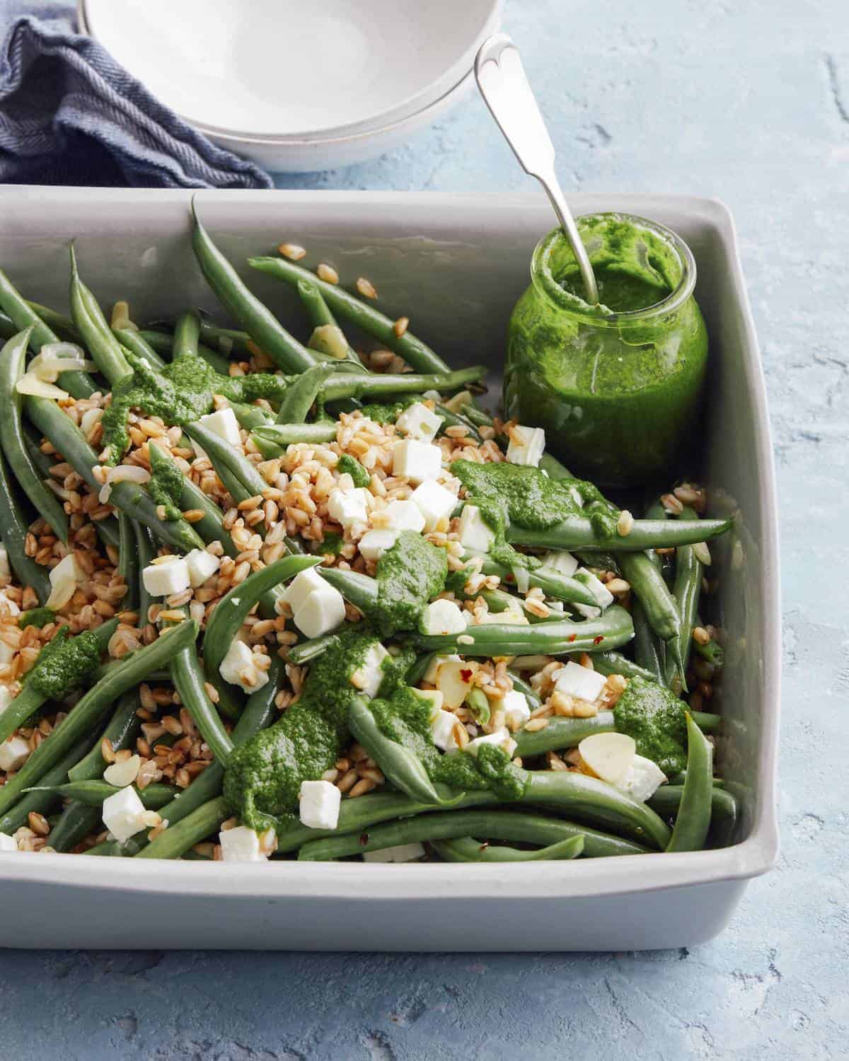 A rectangular dish with green bean farro salad with feta, and a mint basil vinaigrette drizzled on top, with a jar of the vinaigrette on the side.