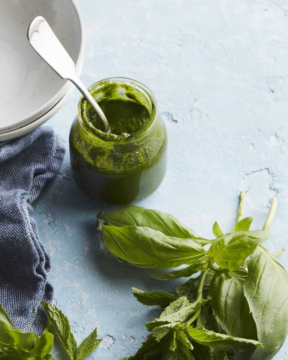 A glass jar with mint basil vinaigrette with a spoon, with some fresh basil, mint, some plates and a kitchen towel on the side.