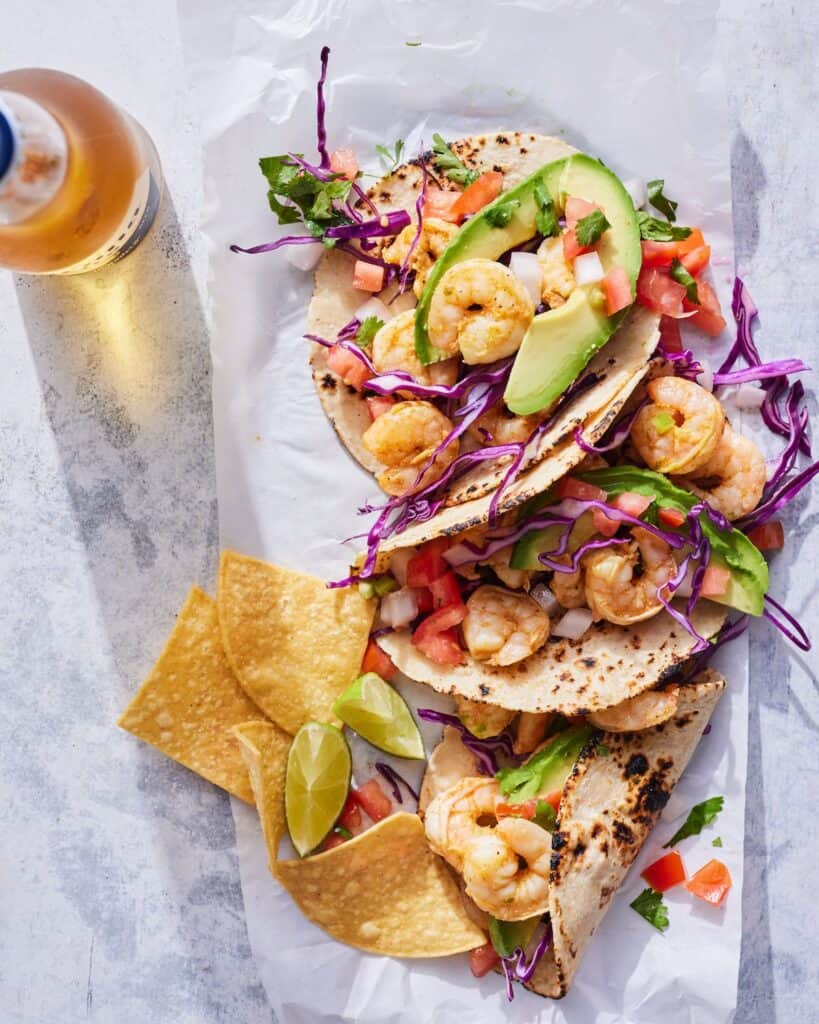 Three shrimp tacos laid out on a counter along with tortilla chips and lime wedges along with a drink on the side.