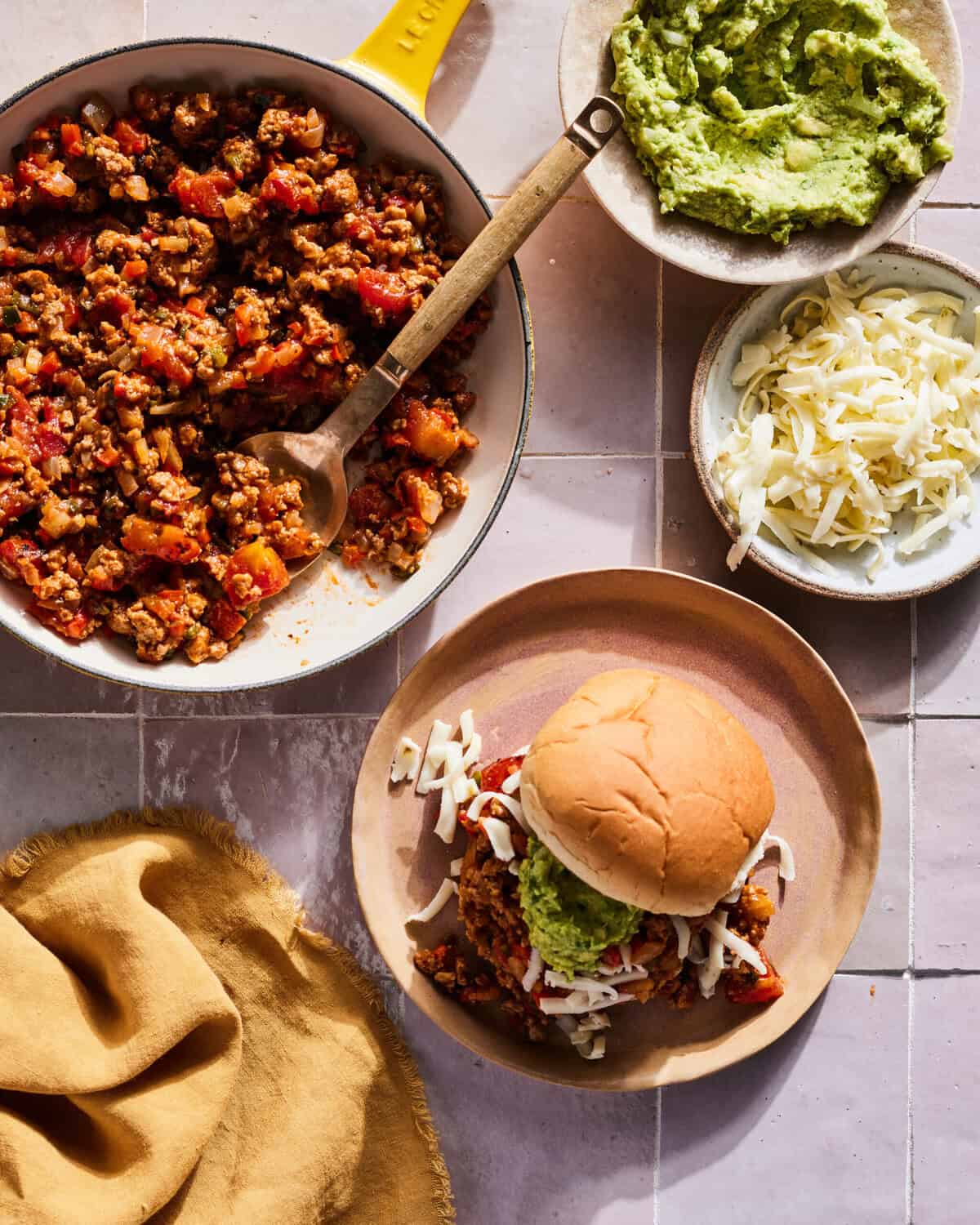 A skillet with cooked ground turkey for a tex mex style sloppy joe, with two bowls on the side with guacamole and shredded cheese, and a plate with a sloppy joe assembled with these ingredients on a bun.