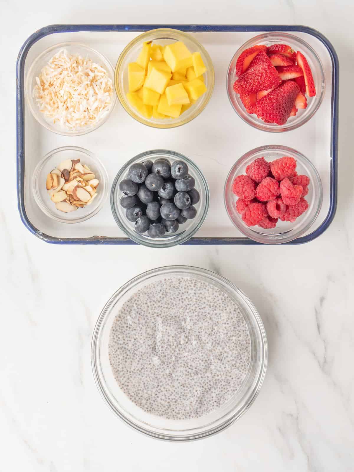 A bowl with chia pudding, and a rectangular platter with six small bowls of toppings, which are mango, strawberry, blueberry, raspberry, sliced almonds and toasted coconut.