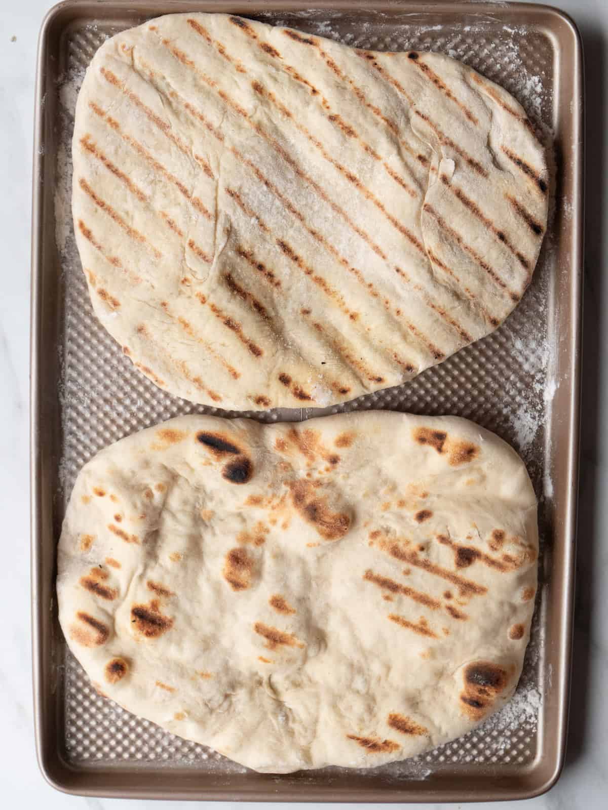 A baking sheet with two freshly grilled pizza base with grill marks.