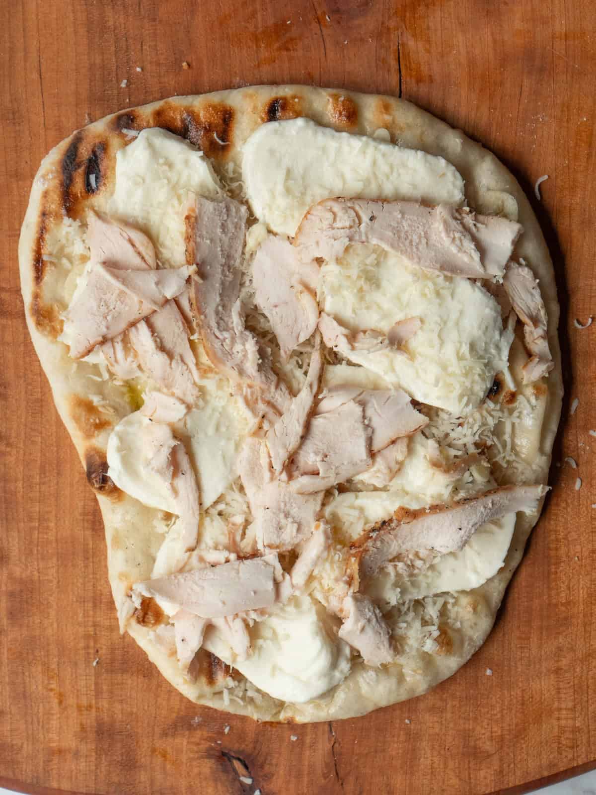 Pizza base with mozzarella, parmesan and chicken breast slices arranged on it ready to be placed back on the grill to melt the cheese.