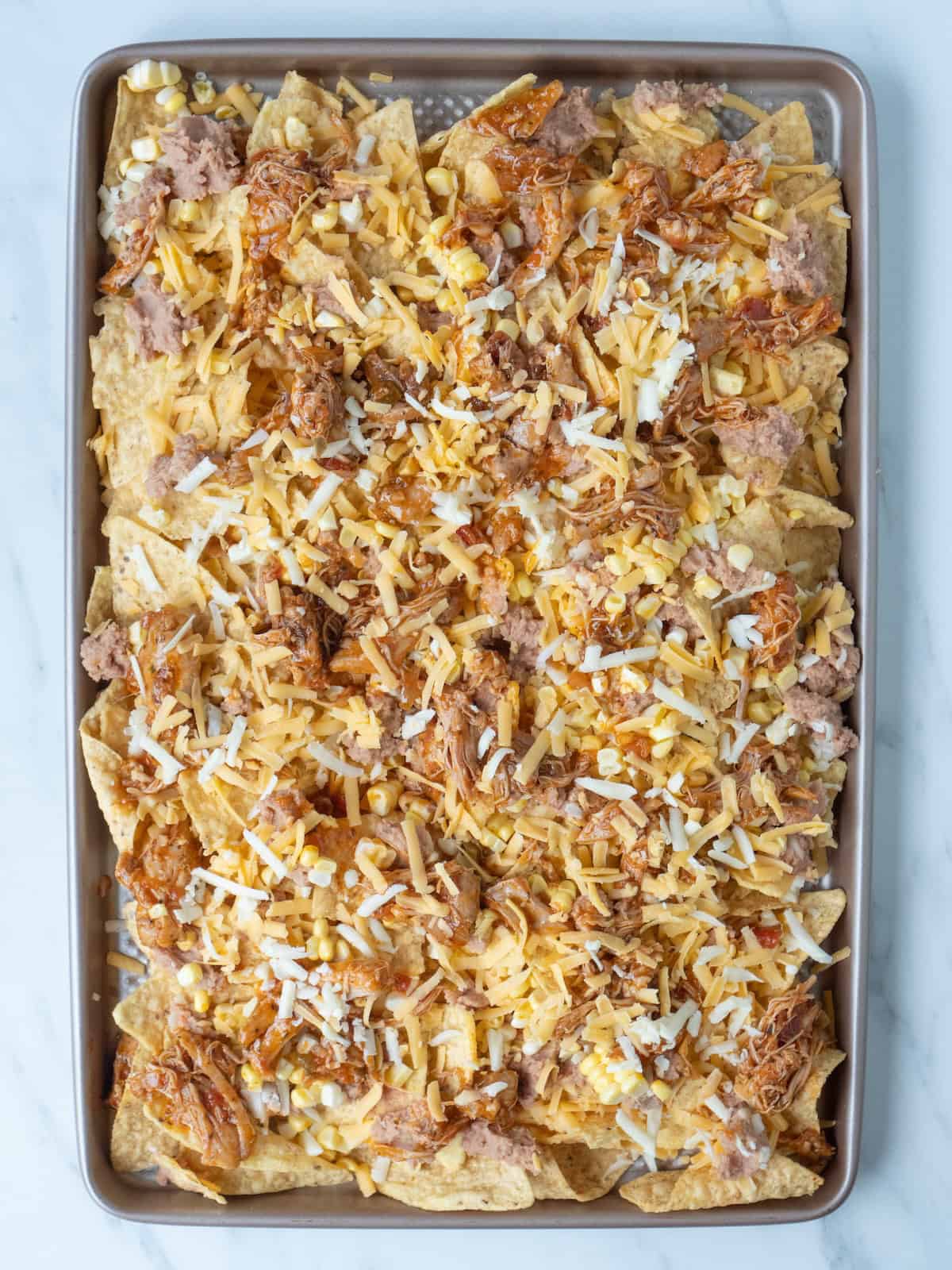 A baking sheet with tortilla chips topped with cheese, refried beans, and chicken evenly.