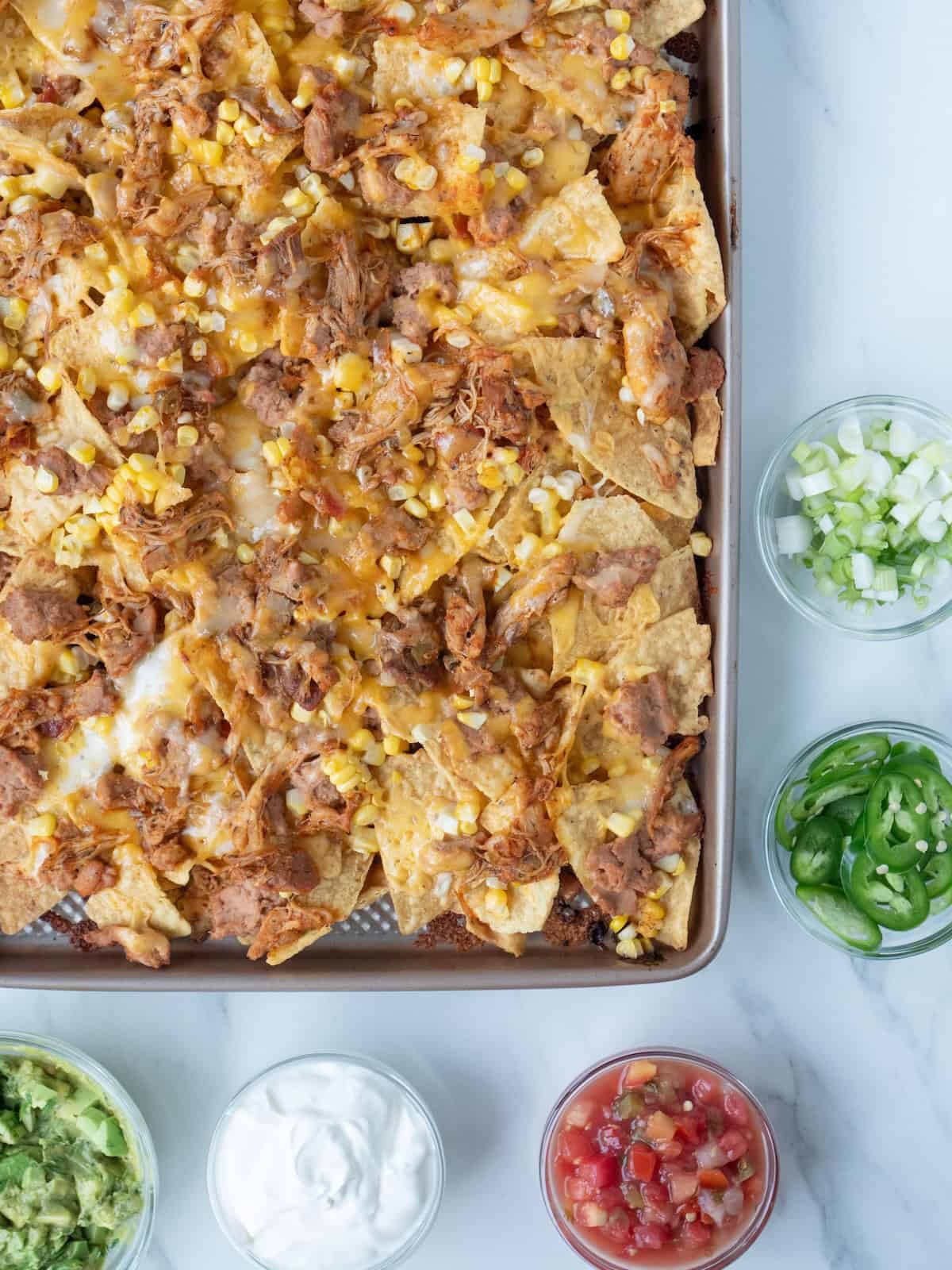 A baking sheet with grilled nachos, along with five small bowls of toppings- guacamole, sour cream, salsa, sliced jalapeños and green onions.