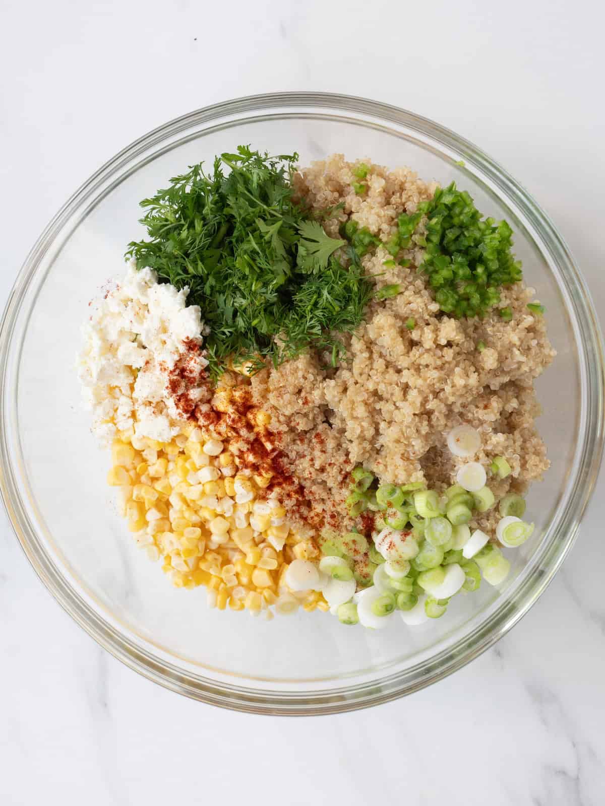 A large glass mixing bowl with corn, green onions, quinoa, cotija cheese, chili powder, cilantro, lime juice and jalapeño to make mexican corn and quinoa salad.