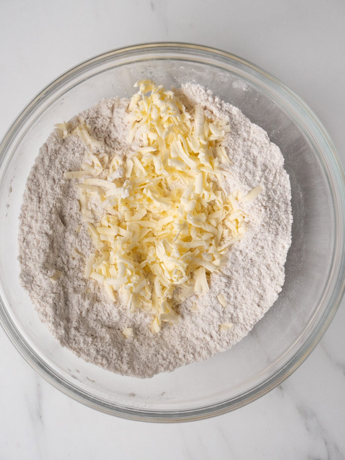 Large glass mixing bowl with grated cold butter added to dry ingredients.
