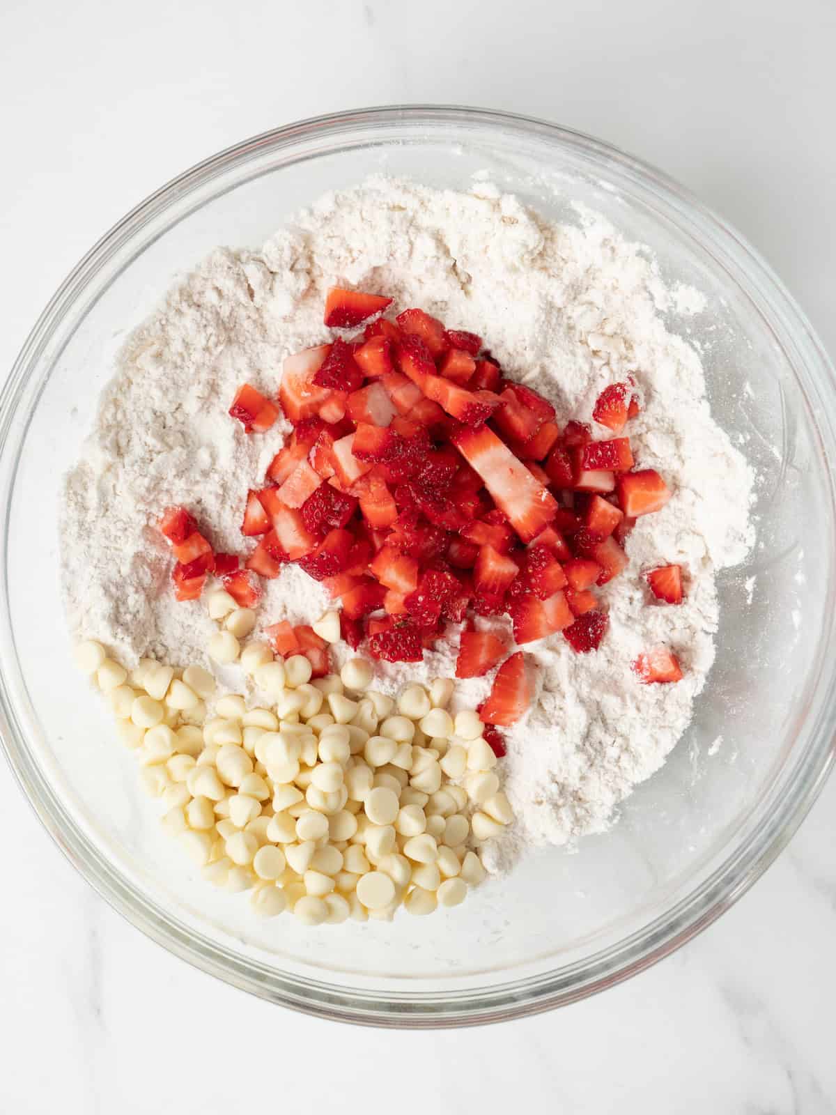 A large glass mixing bowl with white chocolate chips and chopped strawberries added to the flour and butter mixture.