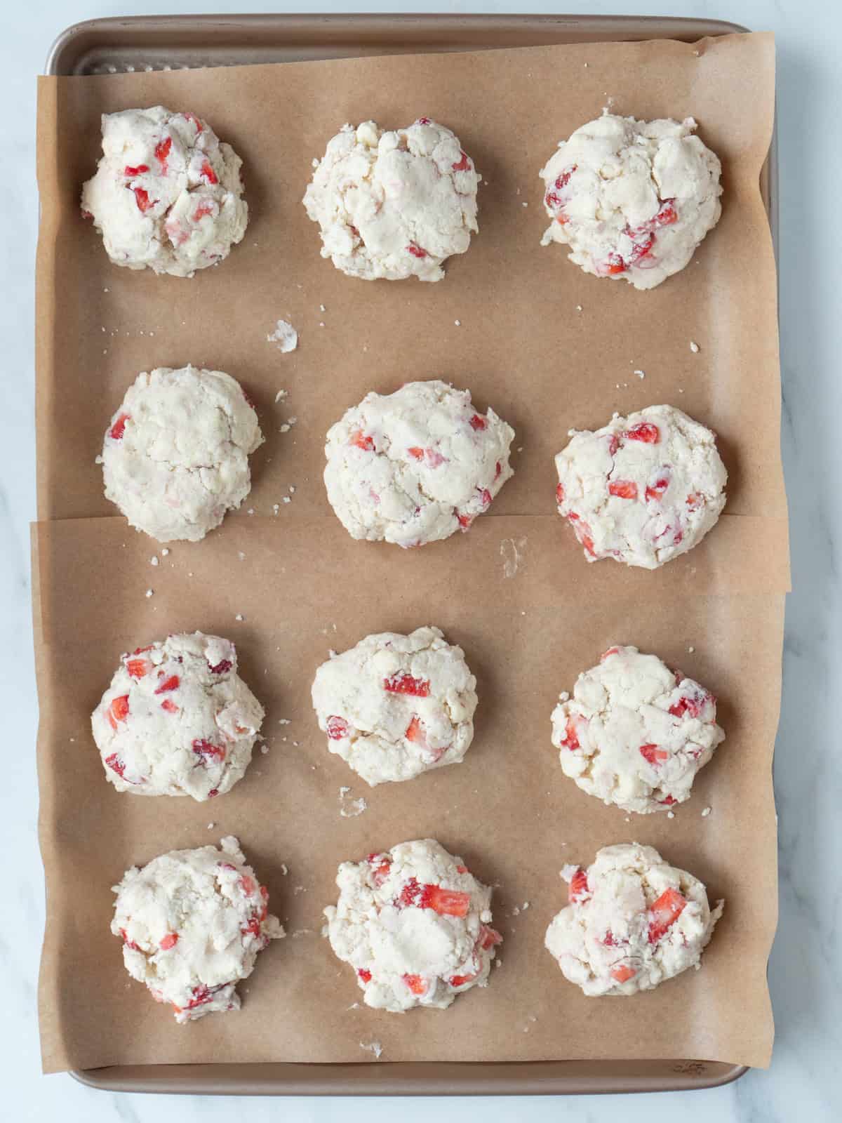 A baking sheet lined with parchment paper and strawberry white chocolate scone dough placed on it as 12 mounds, ready to bake.