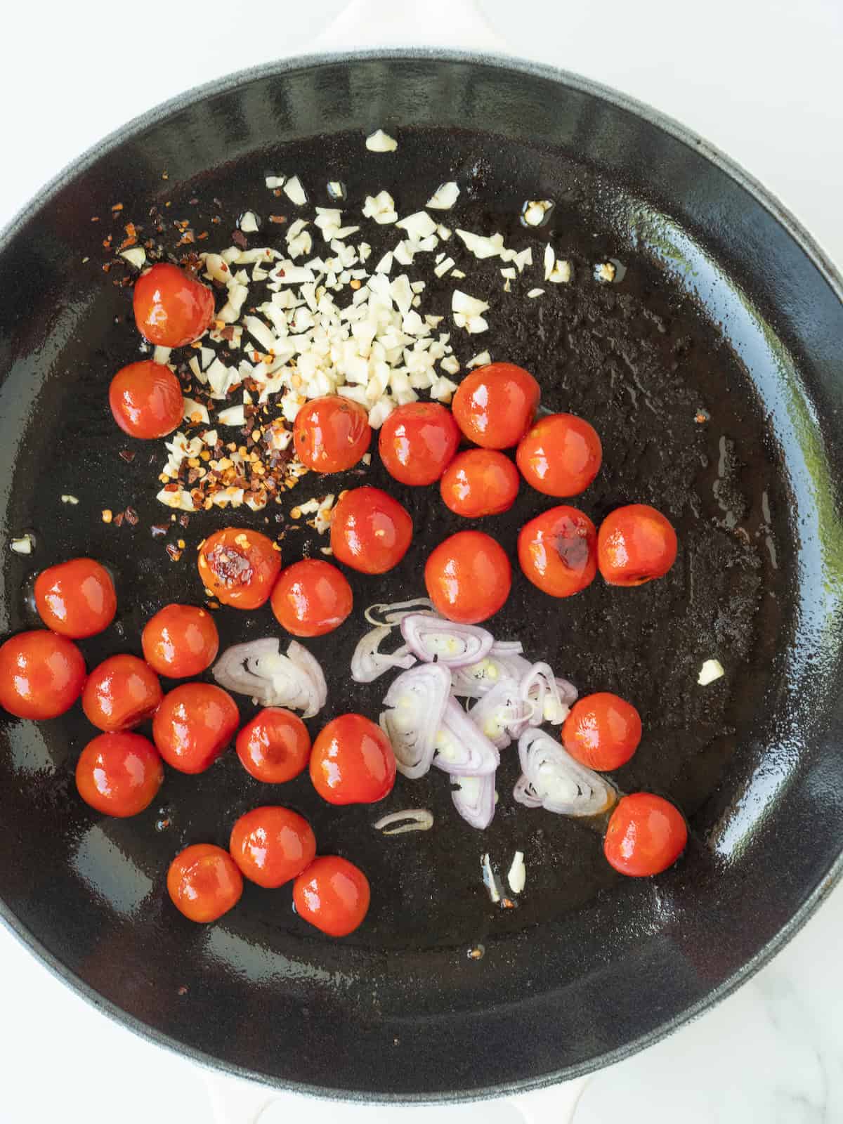 A skillet with cherry tomatoes blistered in olive oil, along with red pepper flakes, shallots and garlic added.