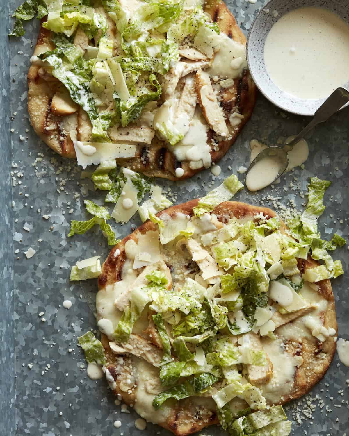 Overhead shot of Chicken Caesar Salad Pizza with a bowl of Caesar Salad Dressing on the side and some drizzled on the pizza.