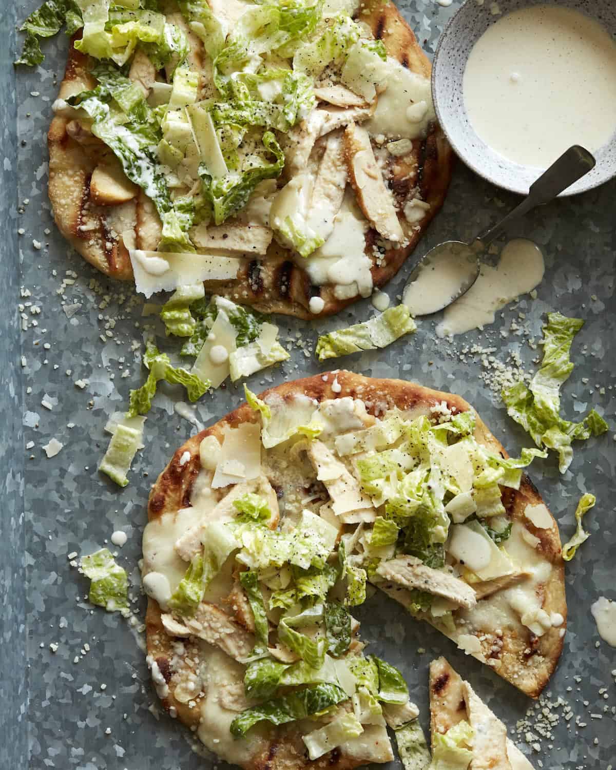 Overhead shot of Chicken Caesar Salad Pizza with a bowl of Caesar Salad Dressing on the side and some drizzled on the pizza.