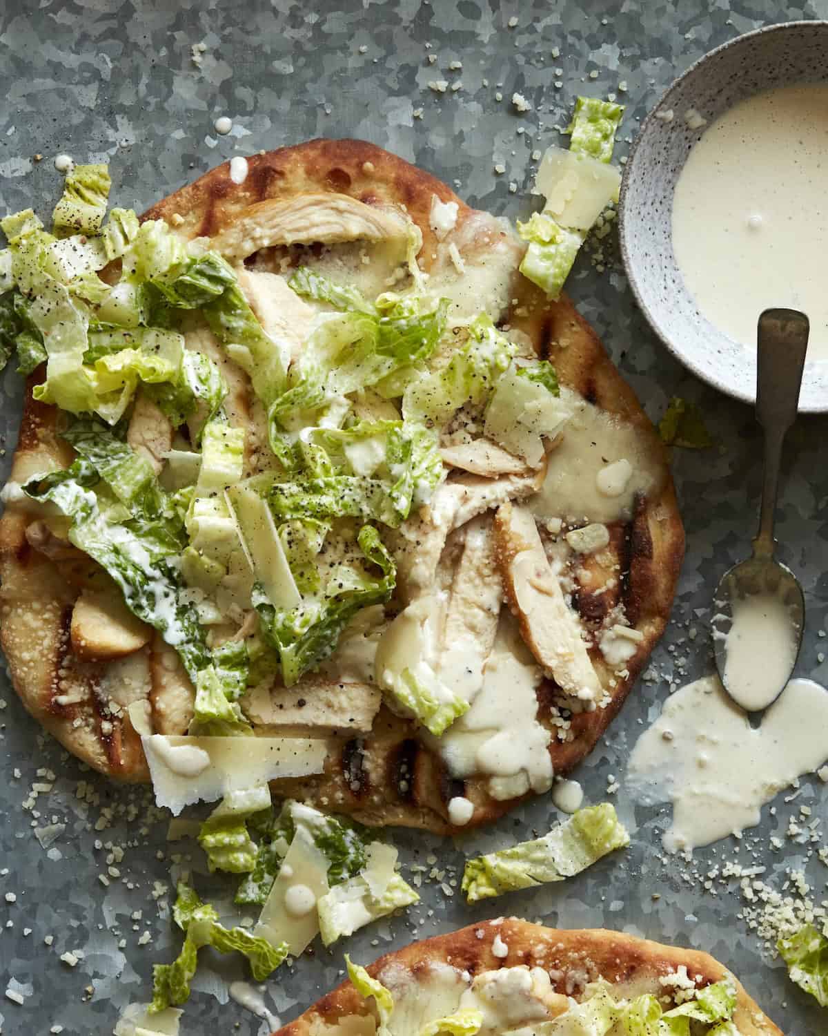 Overhead shot of Chicken Caesar Salad Pizza with a bowl of Caesar Salad Dressing on the side and some drizzled on the pizza.