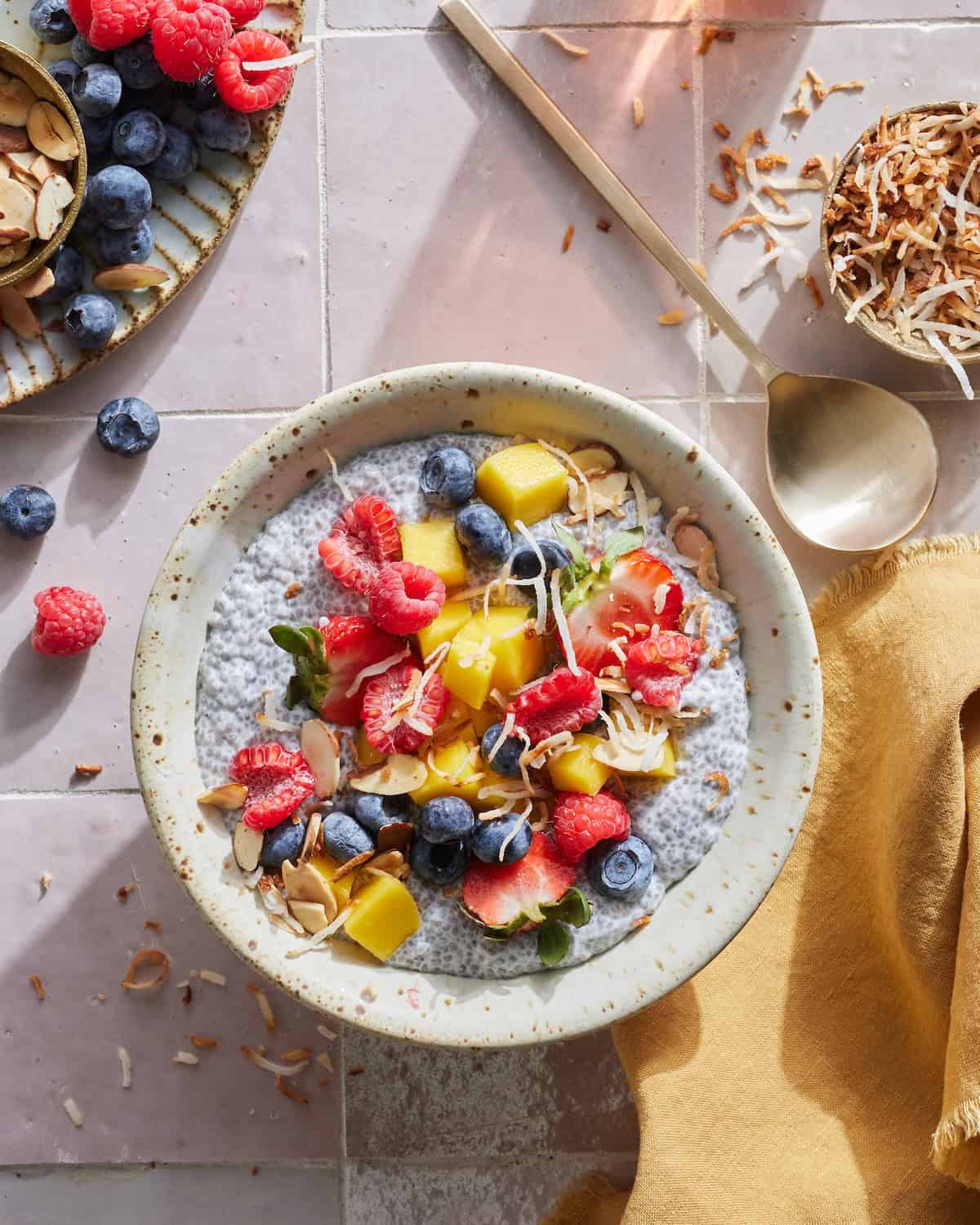 A bowl of chia pudding topped with toasted coconut, sliced almonds, strawberries, raspberries, blueberries and mango with a plate of berries, a bowl of sliced almonds and another bowl of toasted coconut on the side.