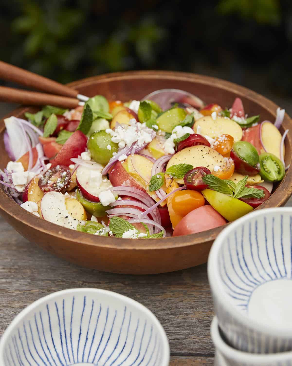 A wooden salad bowl with a stone fruit salad, salad servers and a few bowls on the side for serving.