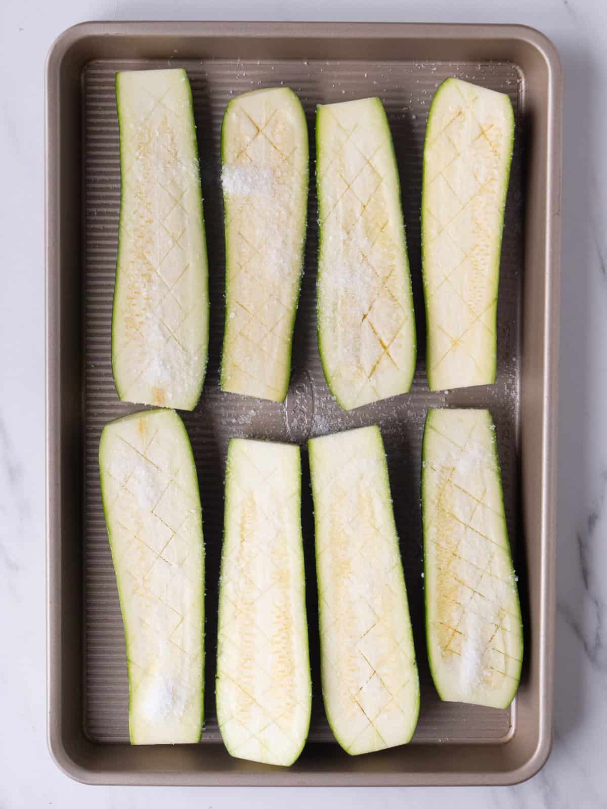 A baking sheet with lengthwise halved zucchini, scored in a crosshatch pattern, and seasoned with salt.