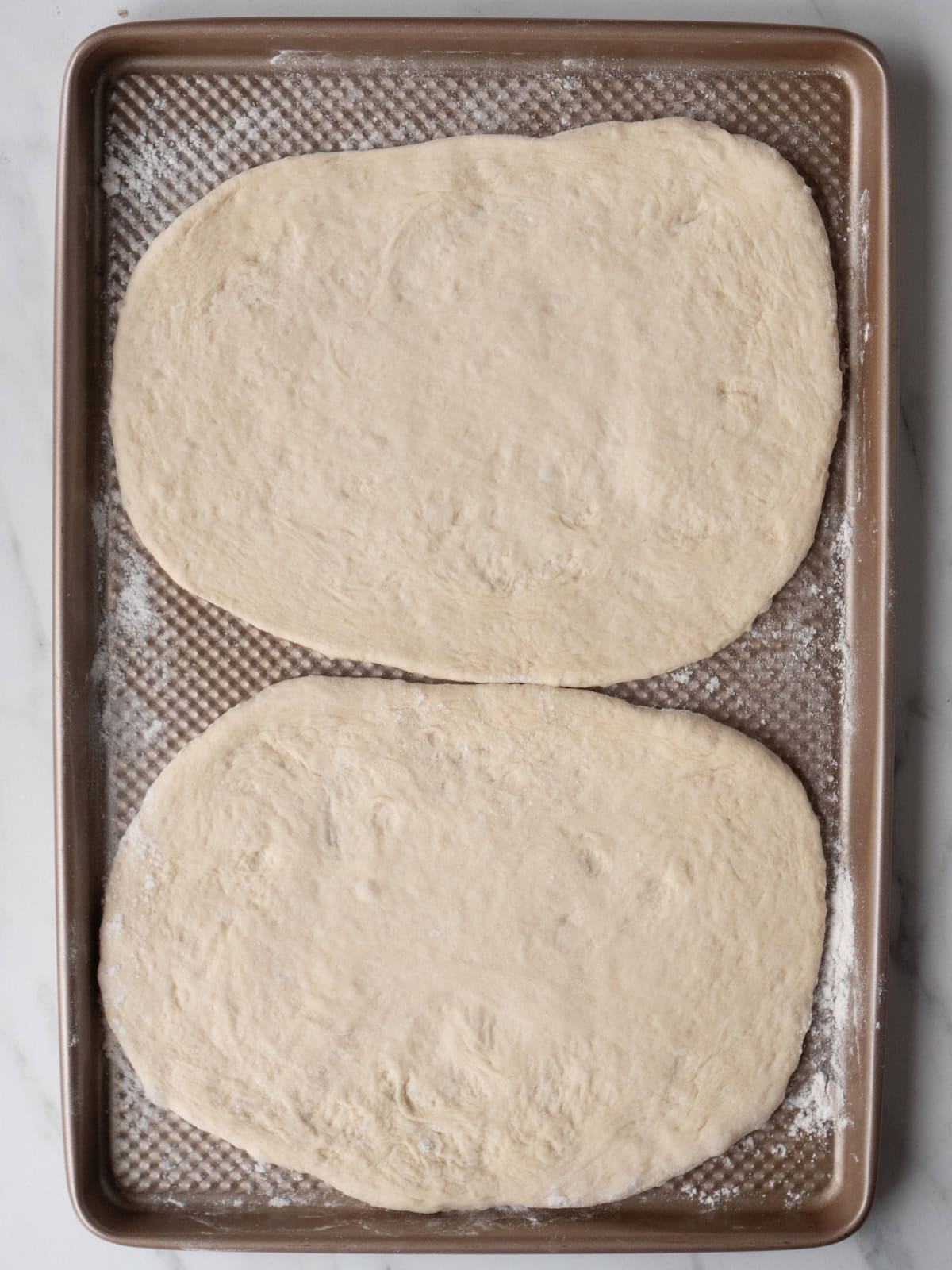 A lightly floured baking sheet with two pizza doughs rolled out into an oval shape ready to be grilled.