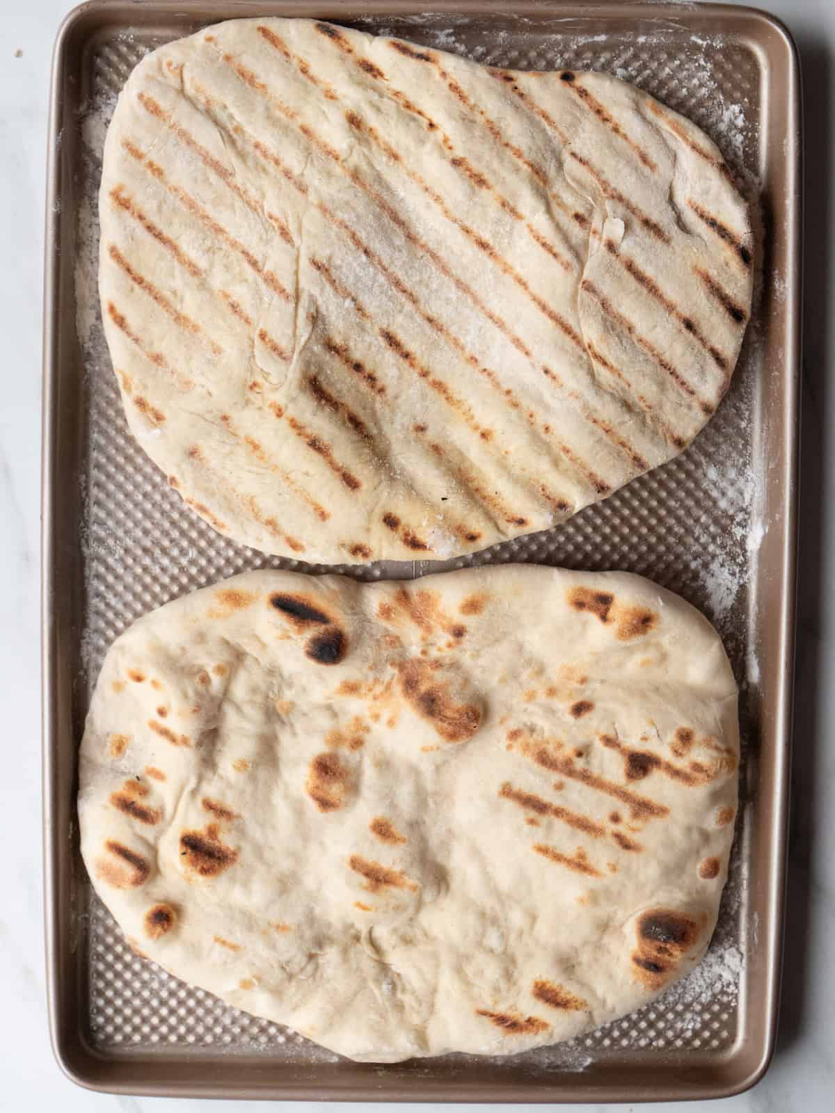 A baking sheet with two pizza dough grilled on a grill with grill marks on both sides.