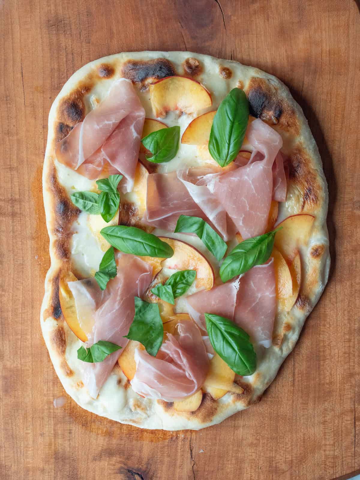 A grilled pizza dough with molten mozzarella taken off the grill add sliced peaches, prosciutto and basil added on top.