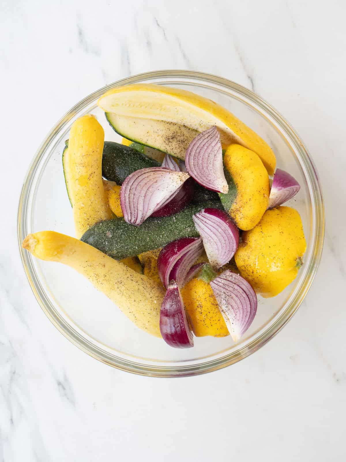 A mixing bowl with patty pan squash, yellow squash, zucchini and red onions sprinkled with salt, black pepper and olive oil.