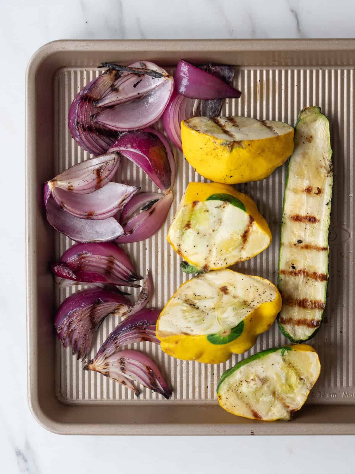 A baking sheet with grilled patty pan squash, yellow squash, zucchini and red onions with grill marks.