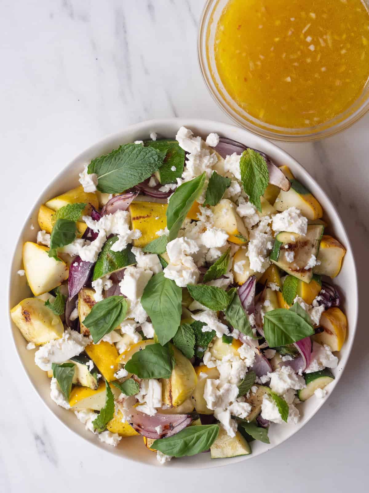 A large bowl of Grilled Summer Squash cubed into pieces topped with Feta, basil leaves and mint leaves along with a bowl of Lemon-Champagne Vinaigrette.