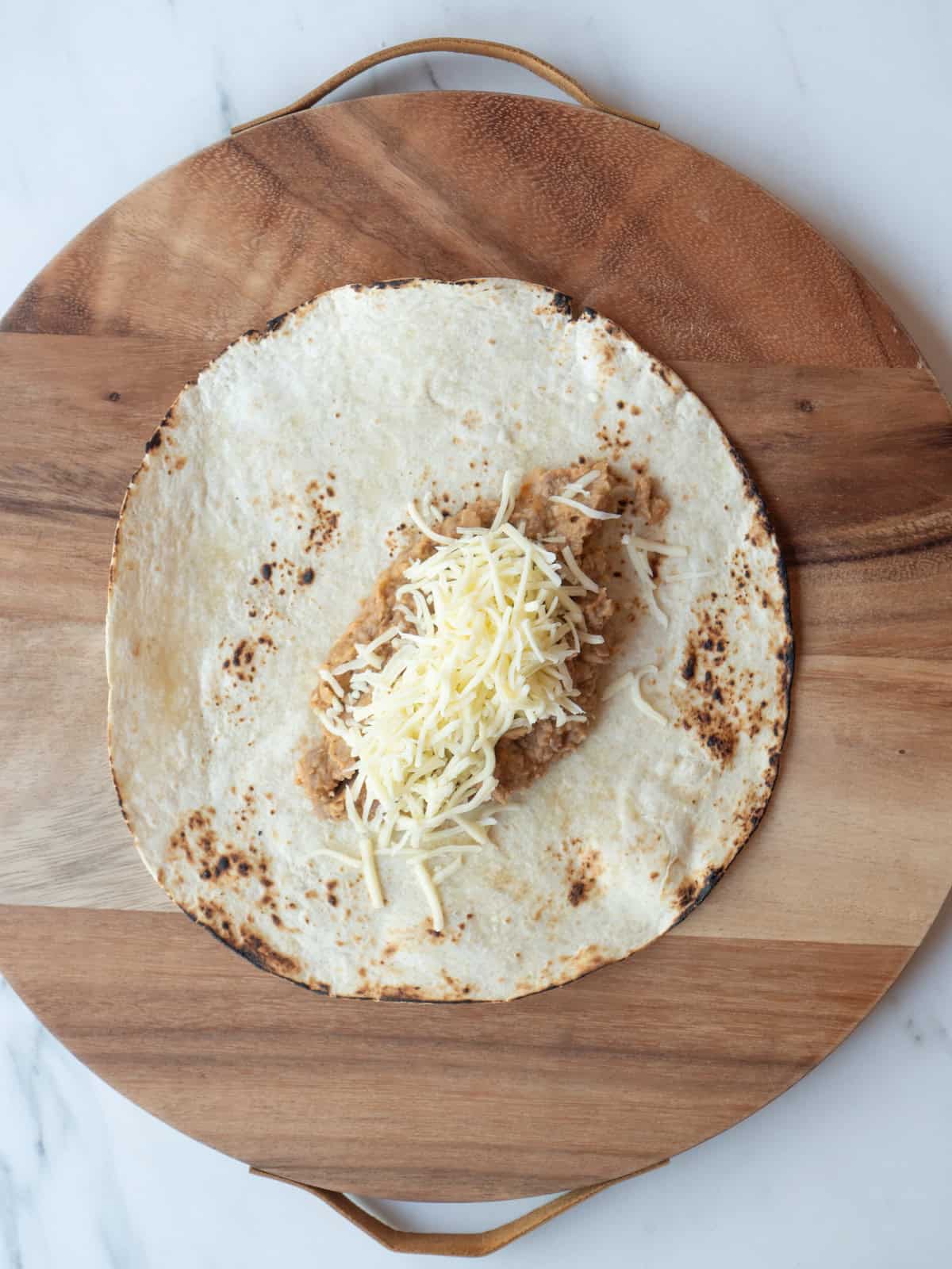 A wooden board with a charred tortilla, topped with refried beans and shredded monterey jack cheese.