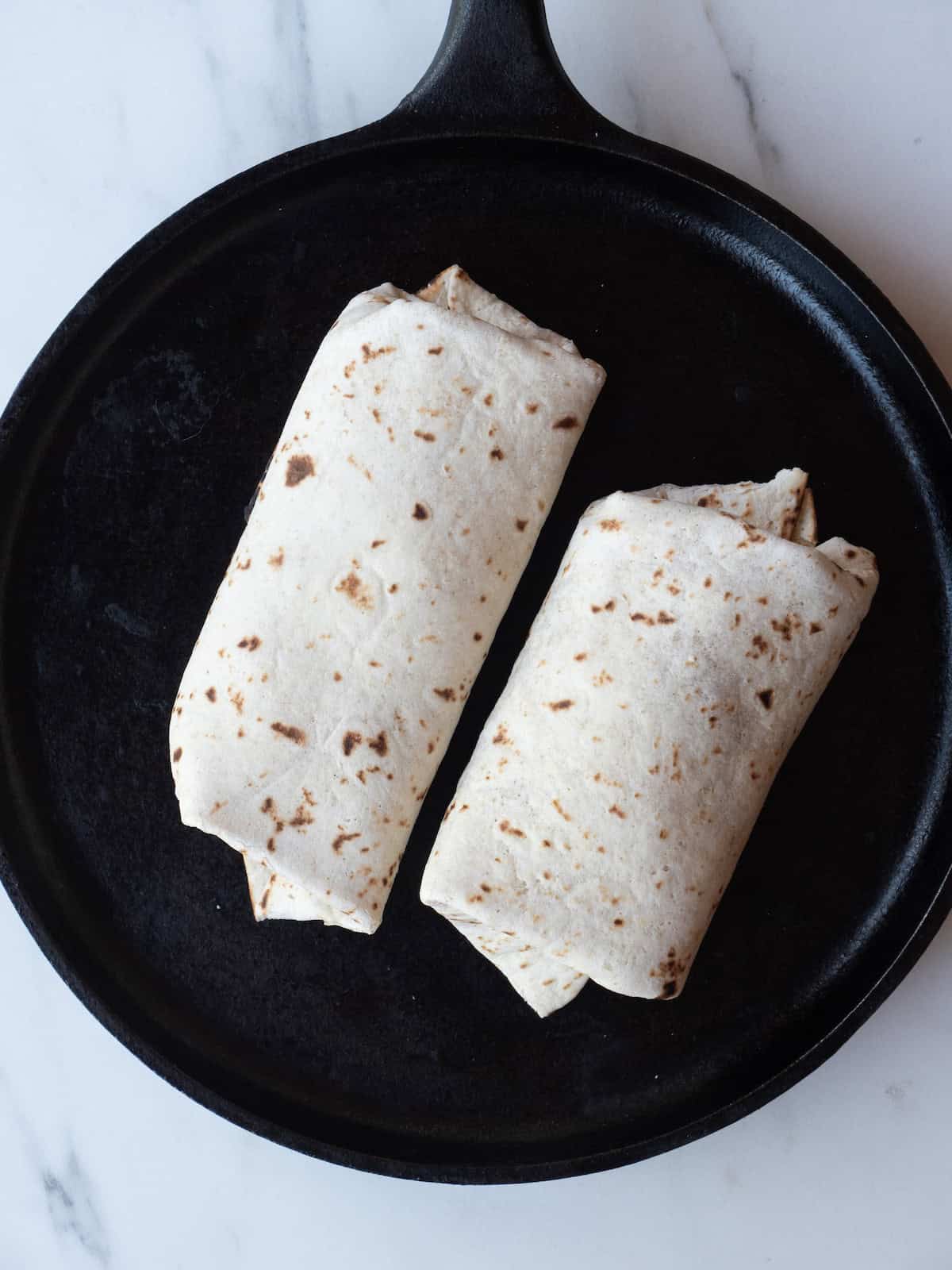 A skillet with two burritos being warmed to give a slight crisp and melt the cheese inside.