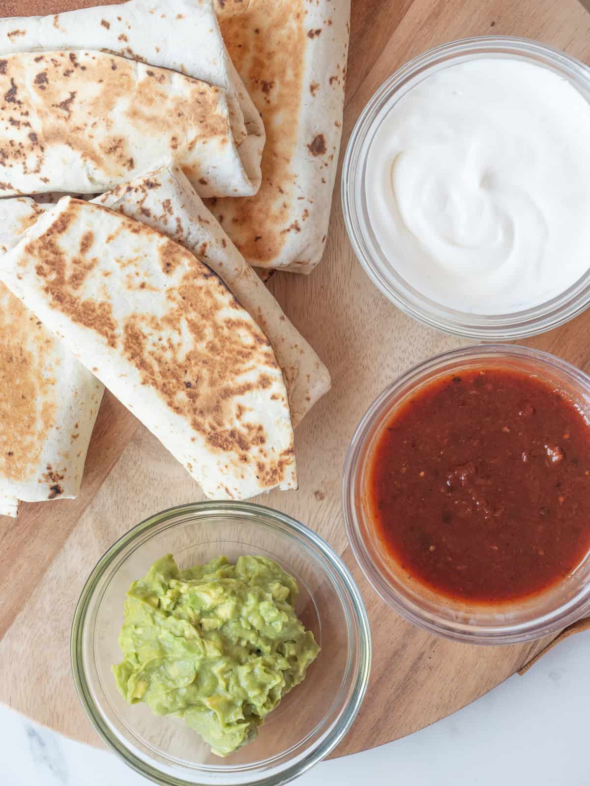 A wooden board with bean and cheese burritos, with three bowls of guacamole, salsa and sour cream.