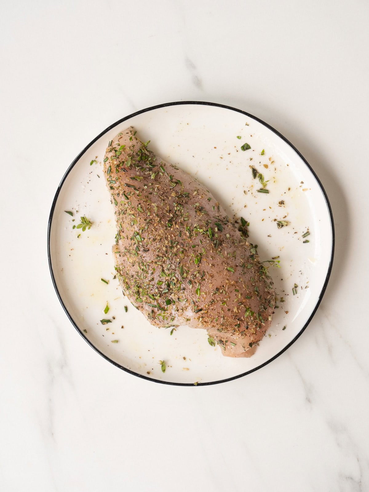 A plate with a chicken breast seasoned with salt and pepper and dried and fresh herbs.