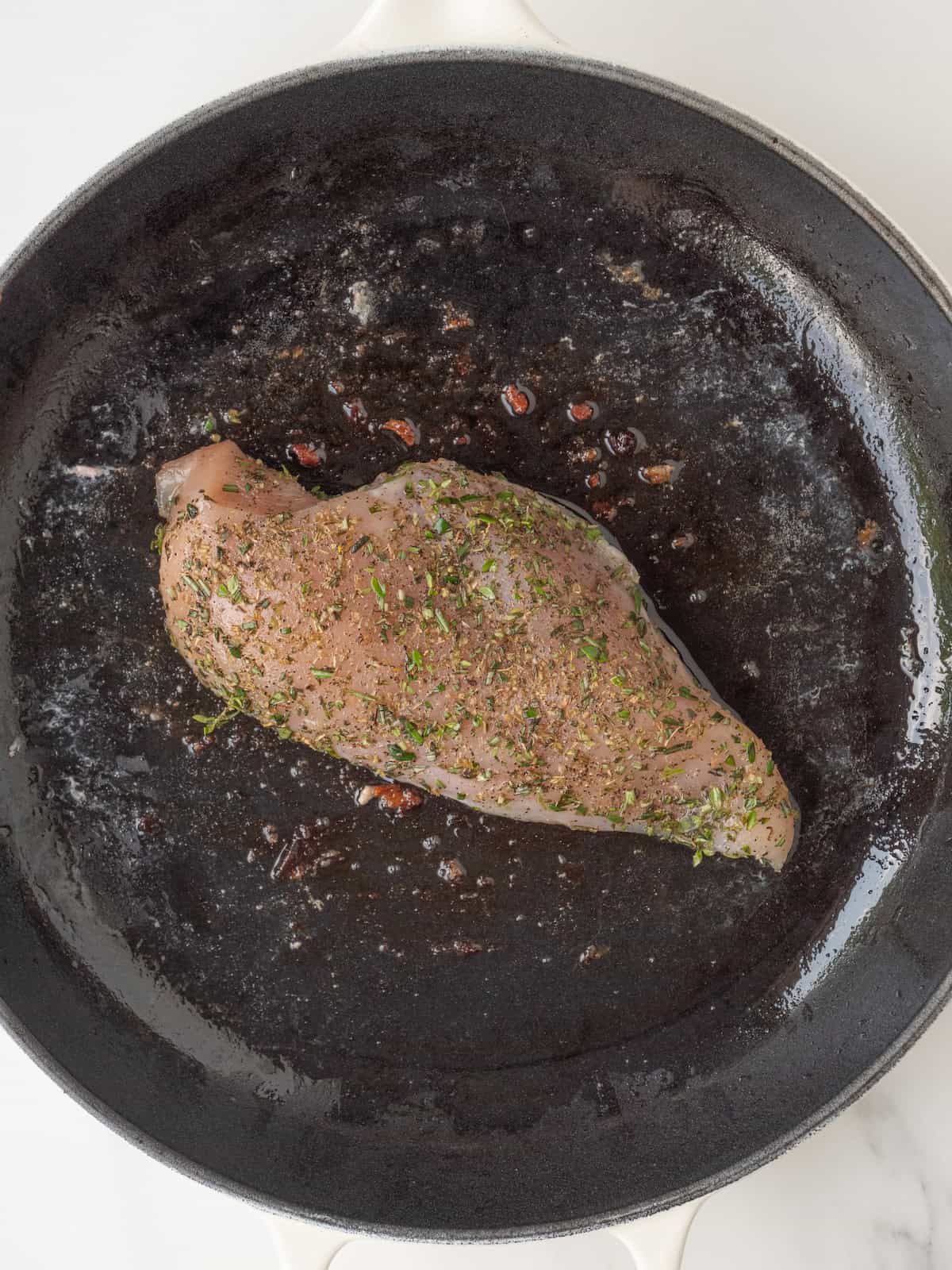 A skillet with a chicken breast seasoned with salt and pepper and dried and fresh herbs, being cooked.