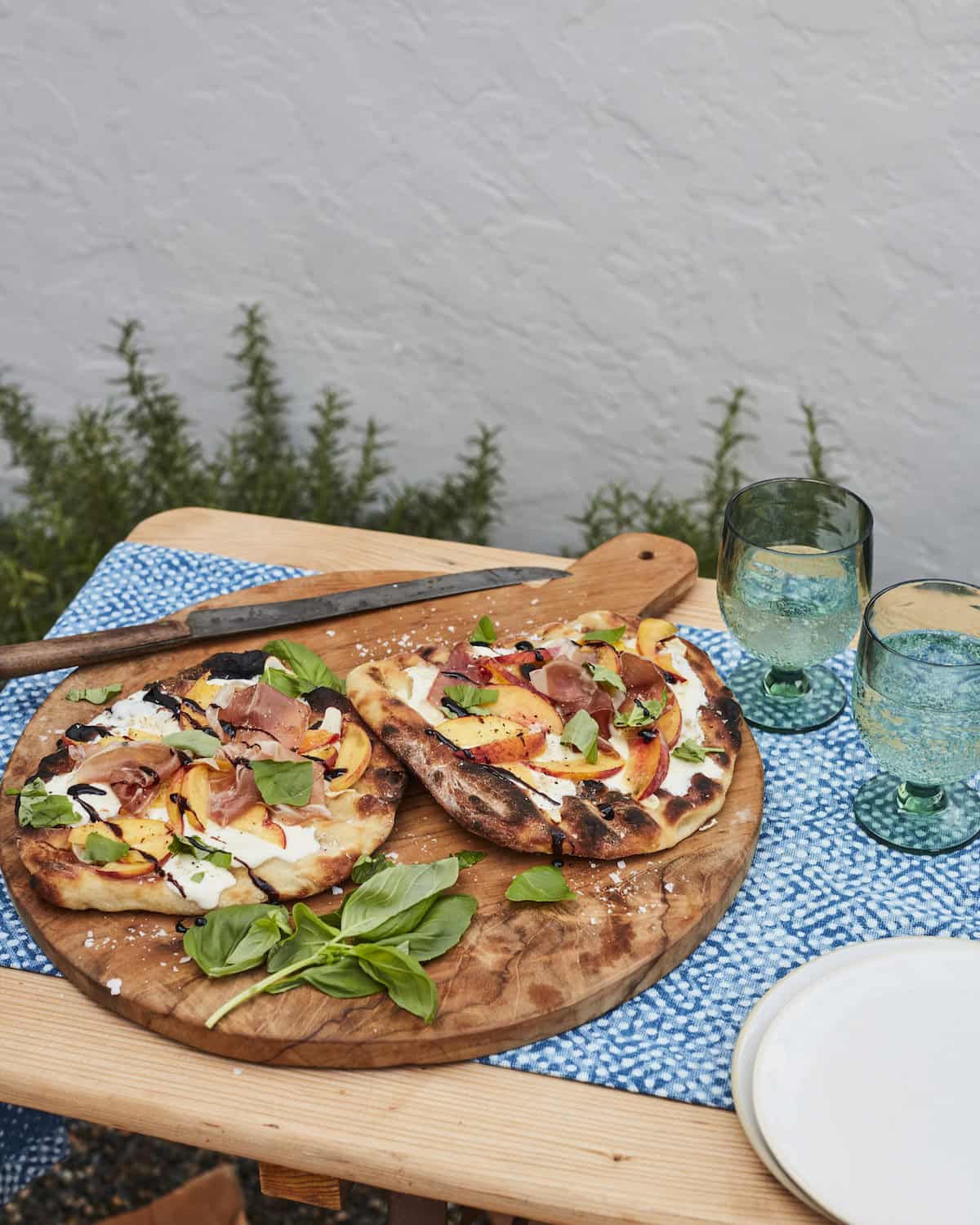 A wooden board with two grilled peach and prosciutto pizzas garnished with basil along with a knife, plates and two fizzy beverages.