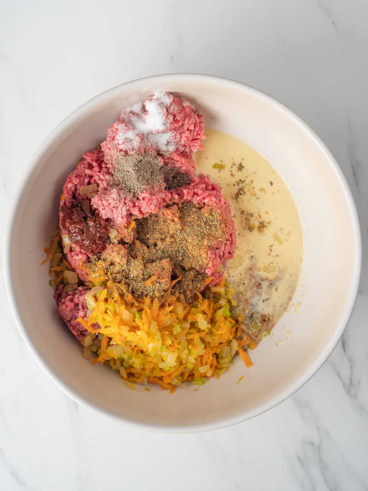 A mixing bowl with an eggs and milk mixture, with ground beef, cooled veggies mixture, Worcestershire sauce, Tuscan seasoning and chipotles added, along with some salt and black pepper.