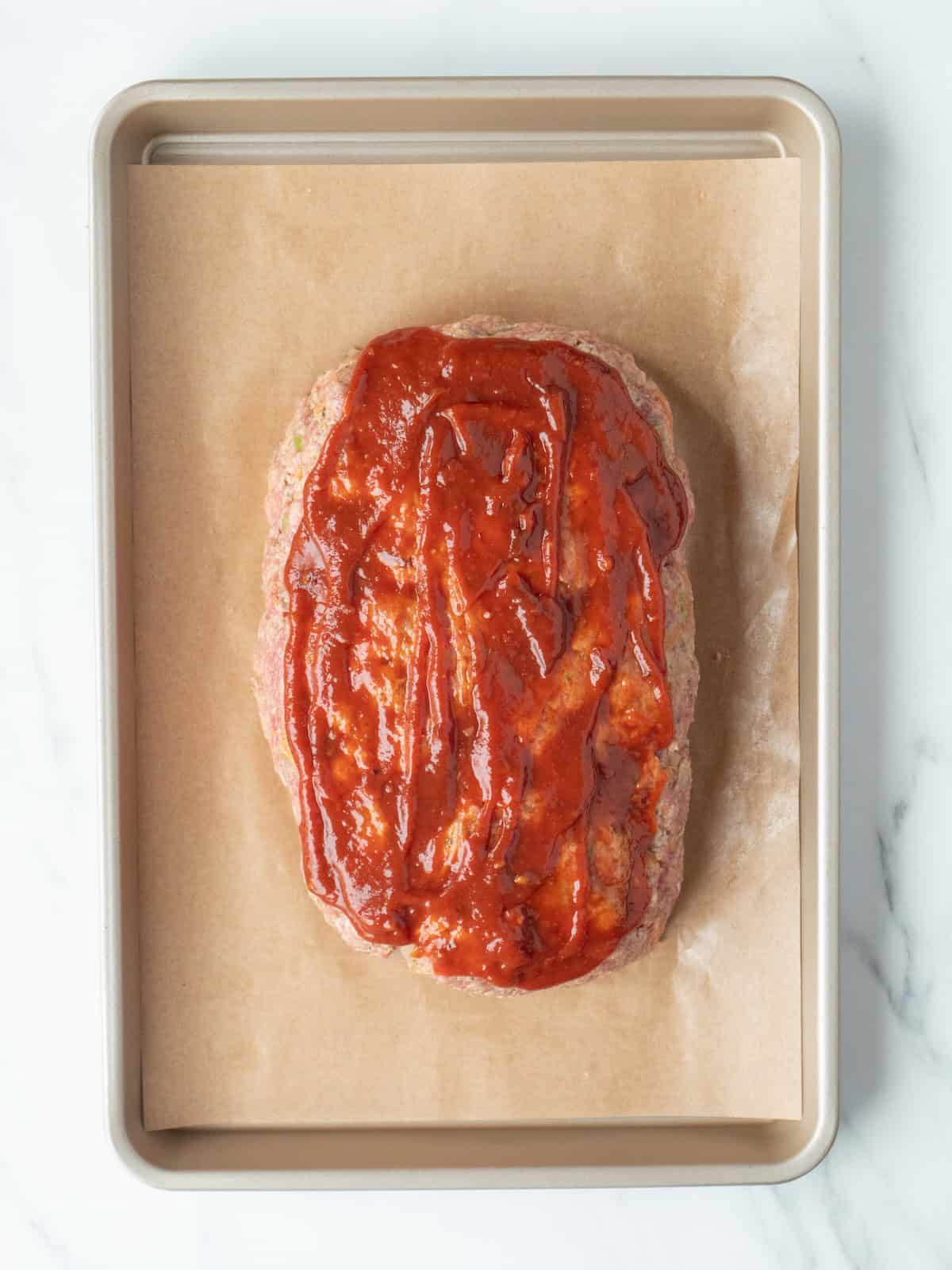 A parchment paper-lined baking sheet with meatloaf mixture shaped into a loaf on it, that has bbq sauce mixture spread evenly on top of the loaf.