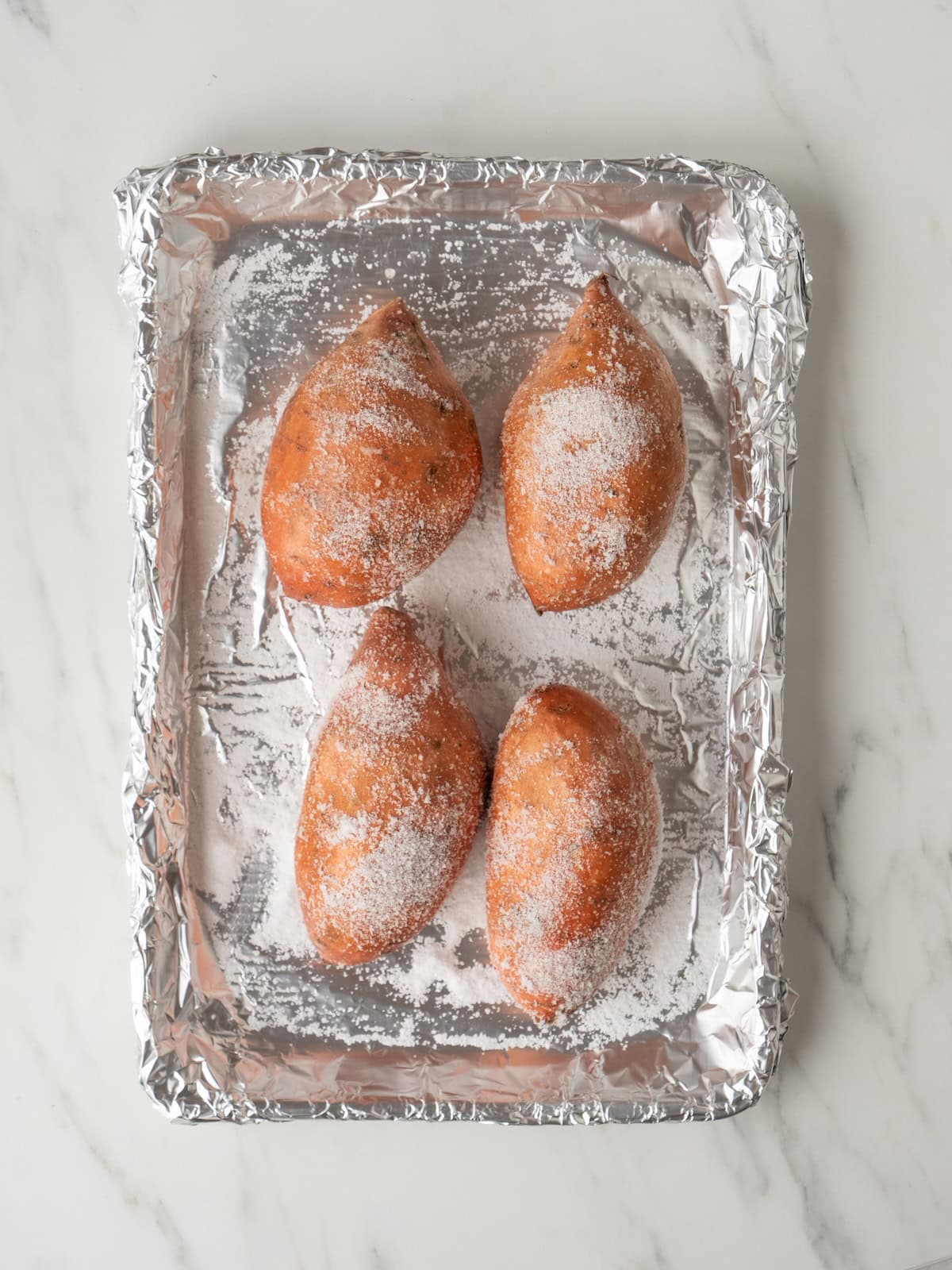 A baking sheet lined with aluminum foil with four sweet potatoes rubbed with kosher salt.