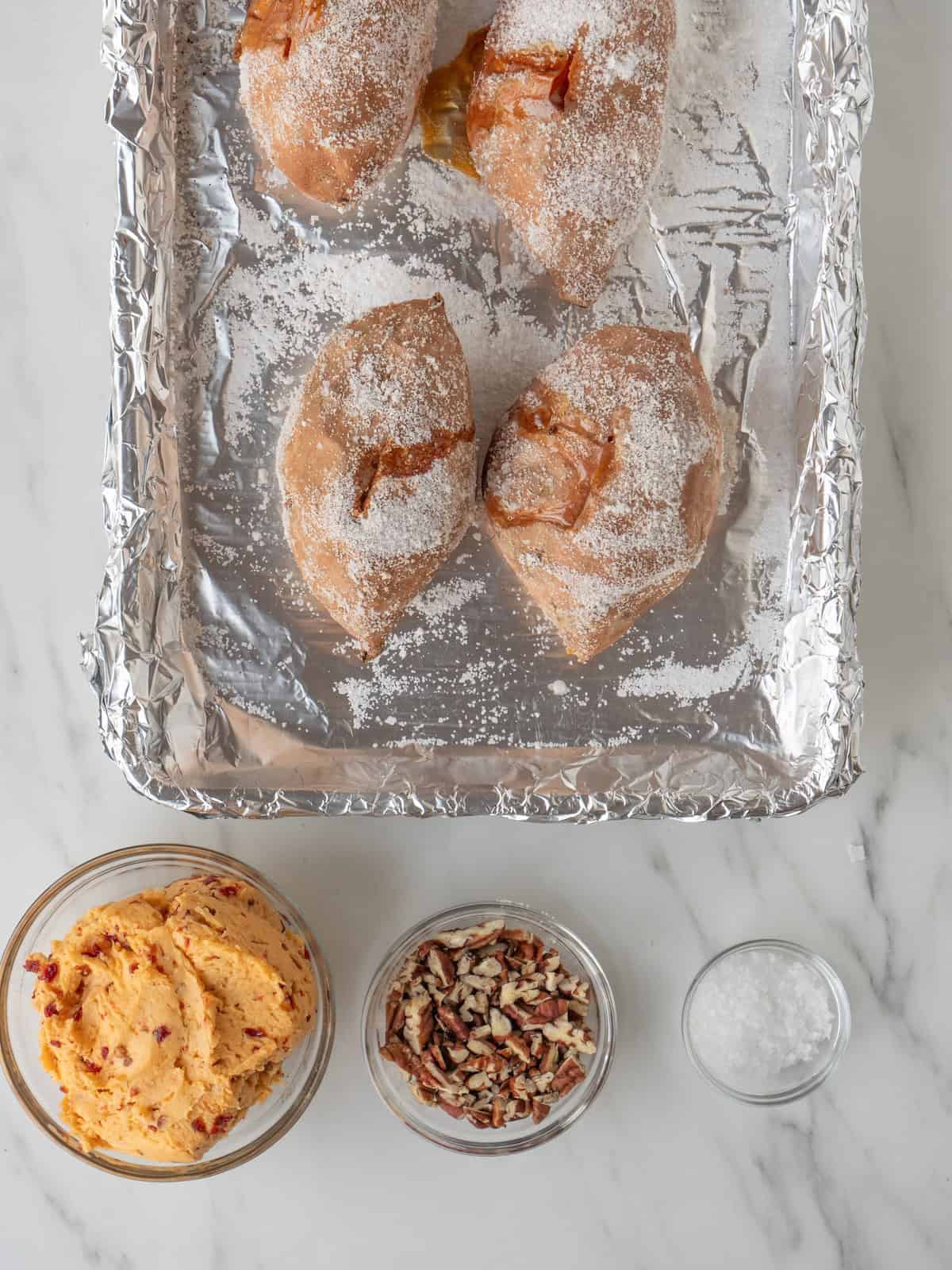 A aluminum foil-lined baking sheet with four sweet potatoes along with three bowls, one of chipotle maple butter, one with pecans and one with salt.