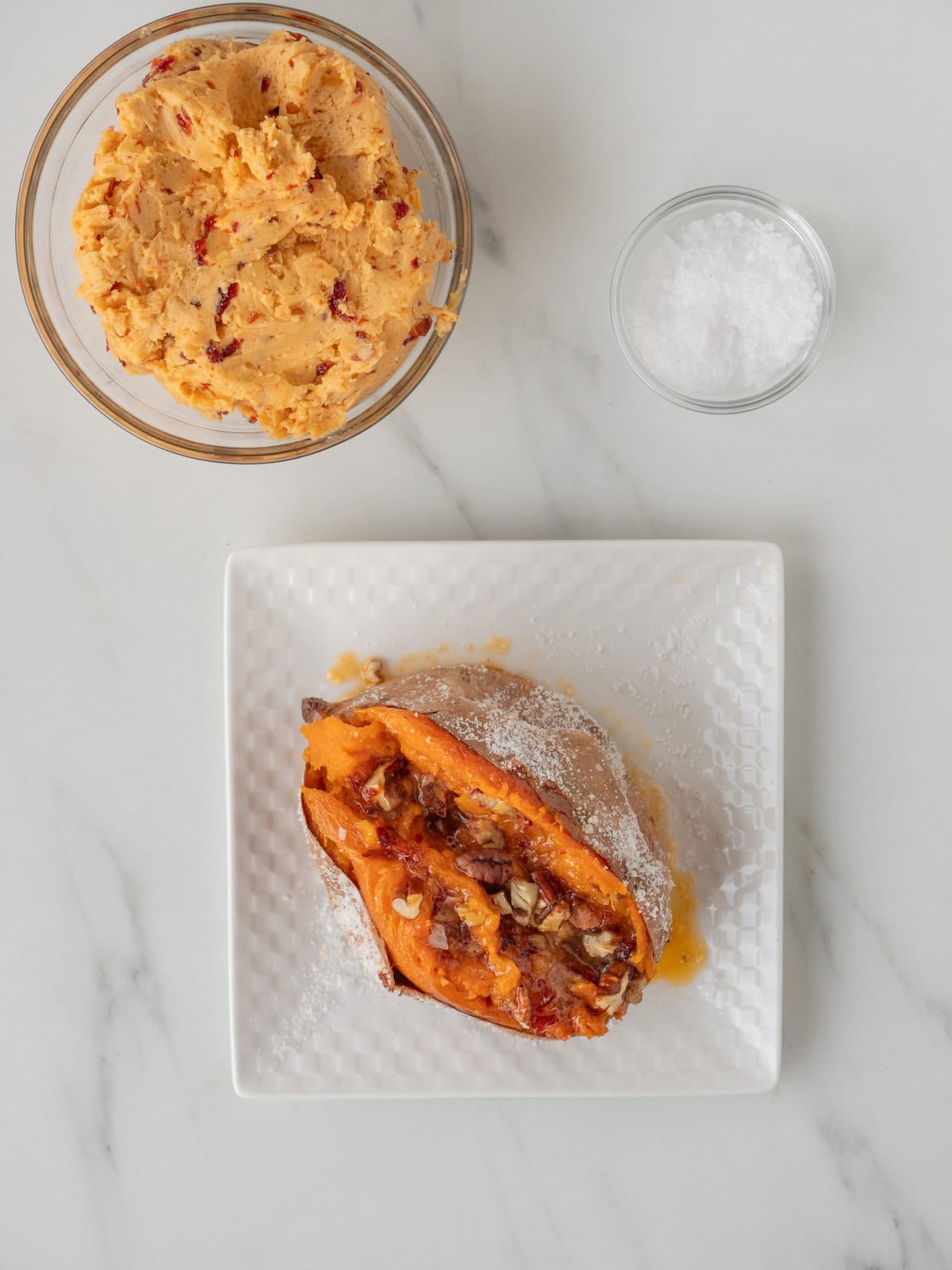 A plate with a gently squeezed baked sweet potato topped with chipotle maple butter, pecans and salt along with a bowl of chipotle maple butter and a small bowl of salt.