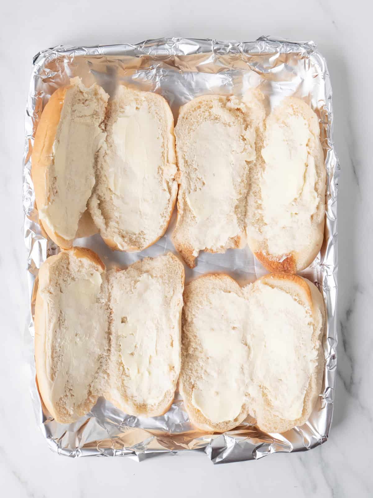 A baking sheet wrapped with foil cut lengthwise, with butter spread on them and placed open-faced, butter side up ready to be toasted.