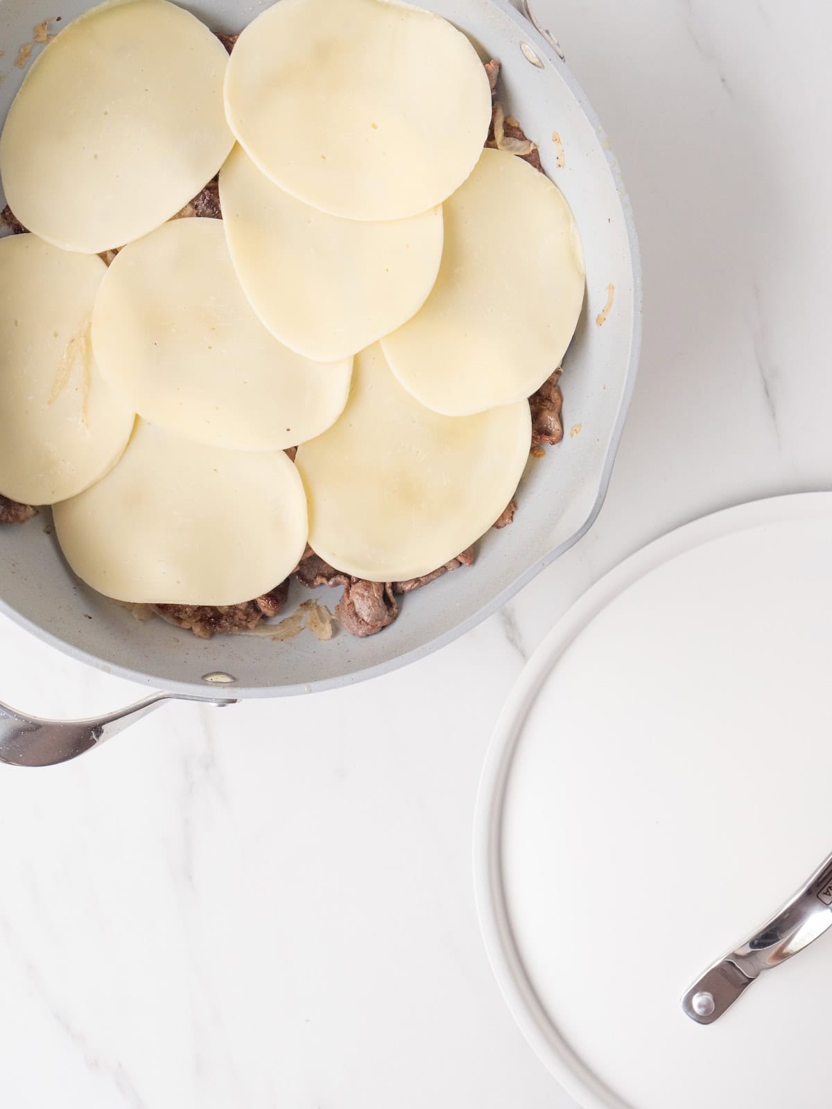 A skillet with cooked onions and steak, spread in a flat layer and topped with provolone cheese slices to let the cheese melt.