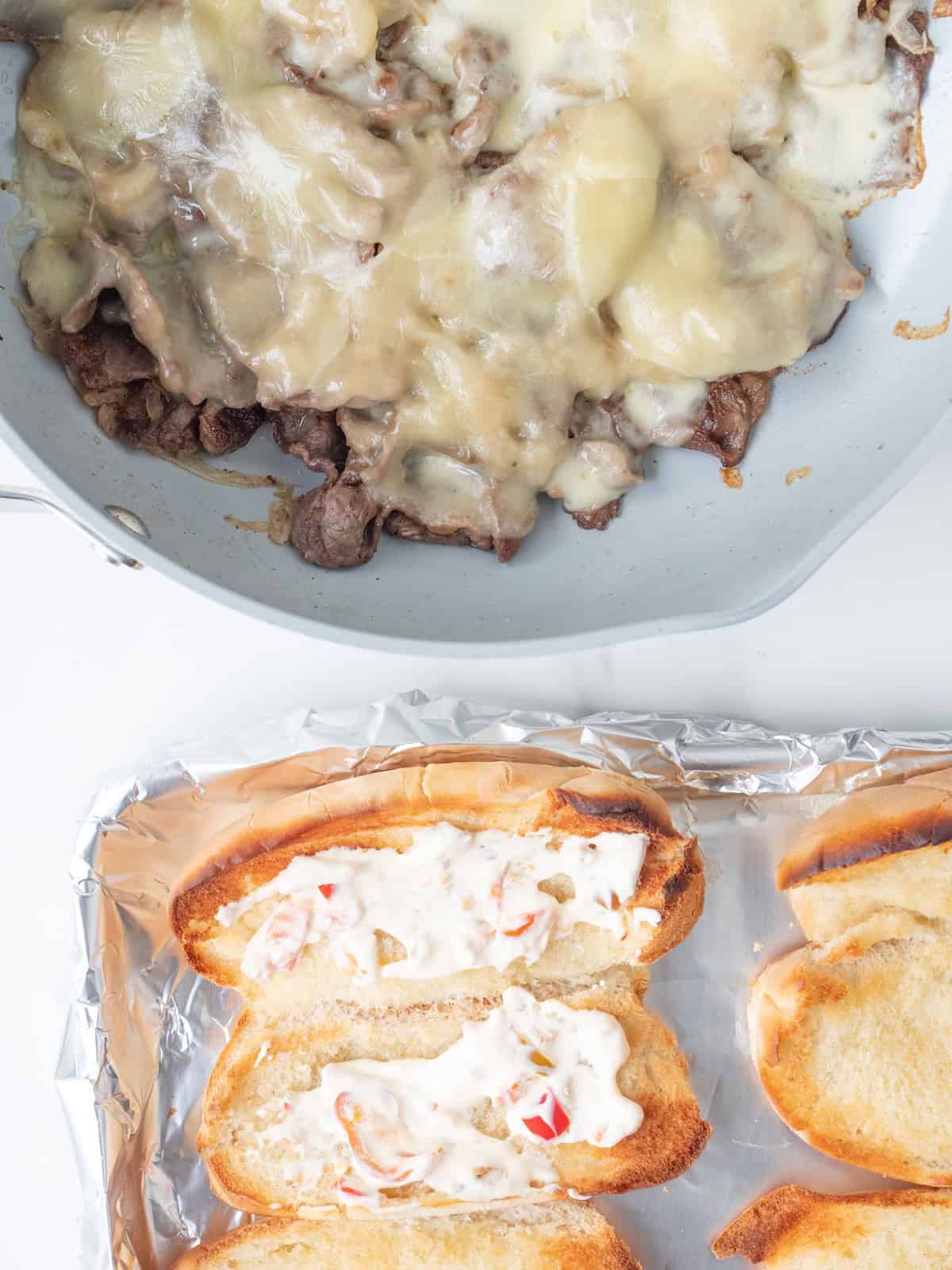 A skillet with cooked onions and steak with melted cheese on top, along with a baking sheet of toasted hoagie rolls with cherry pepper mayonnaise spread on them.