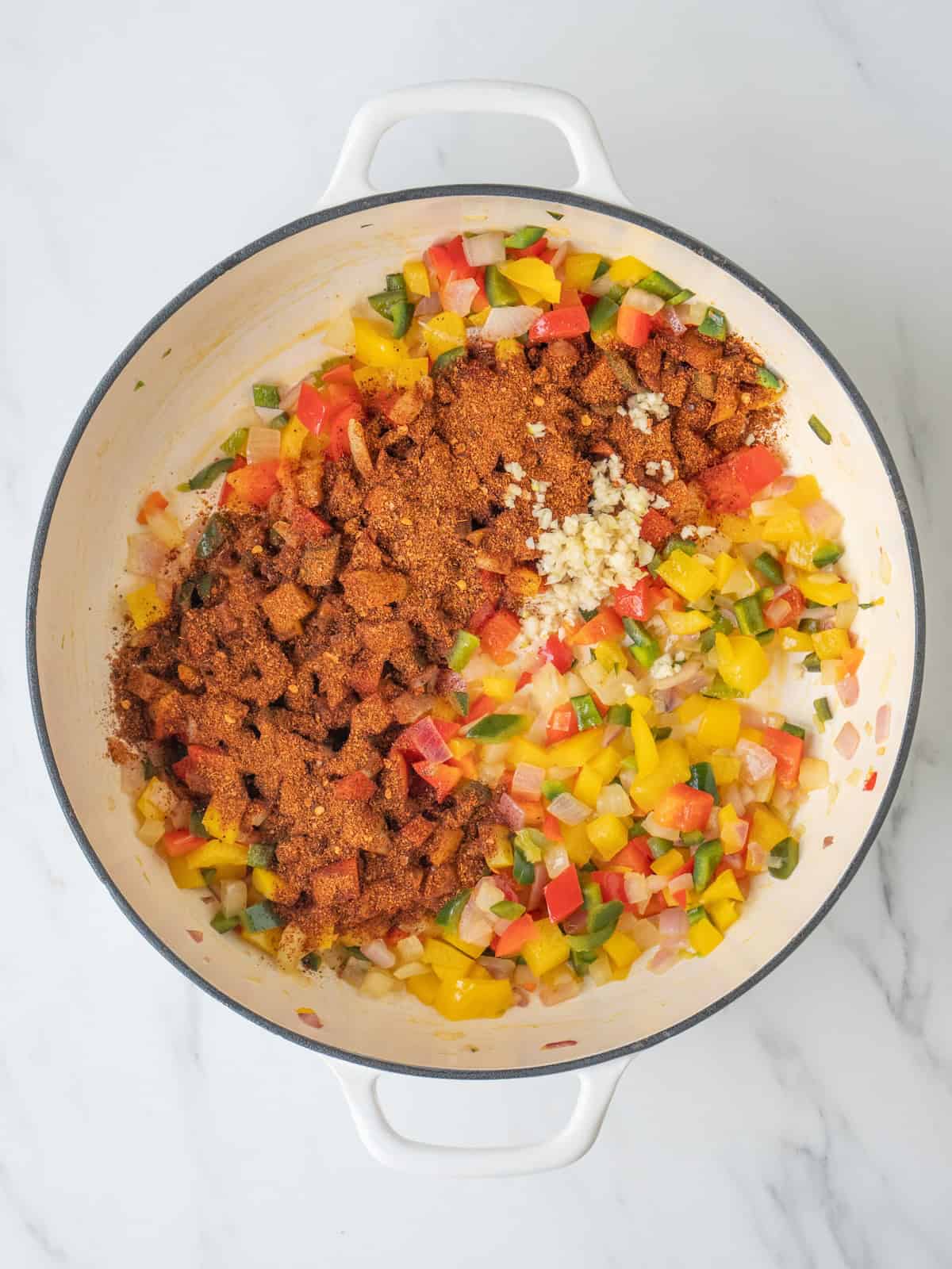 A dutch oven with bell peppers, onions and poblano pepper being sautéed and red chili and garlic added to it.