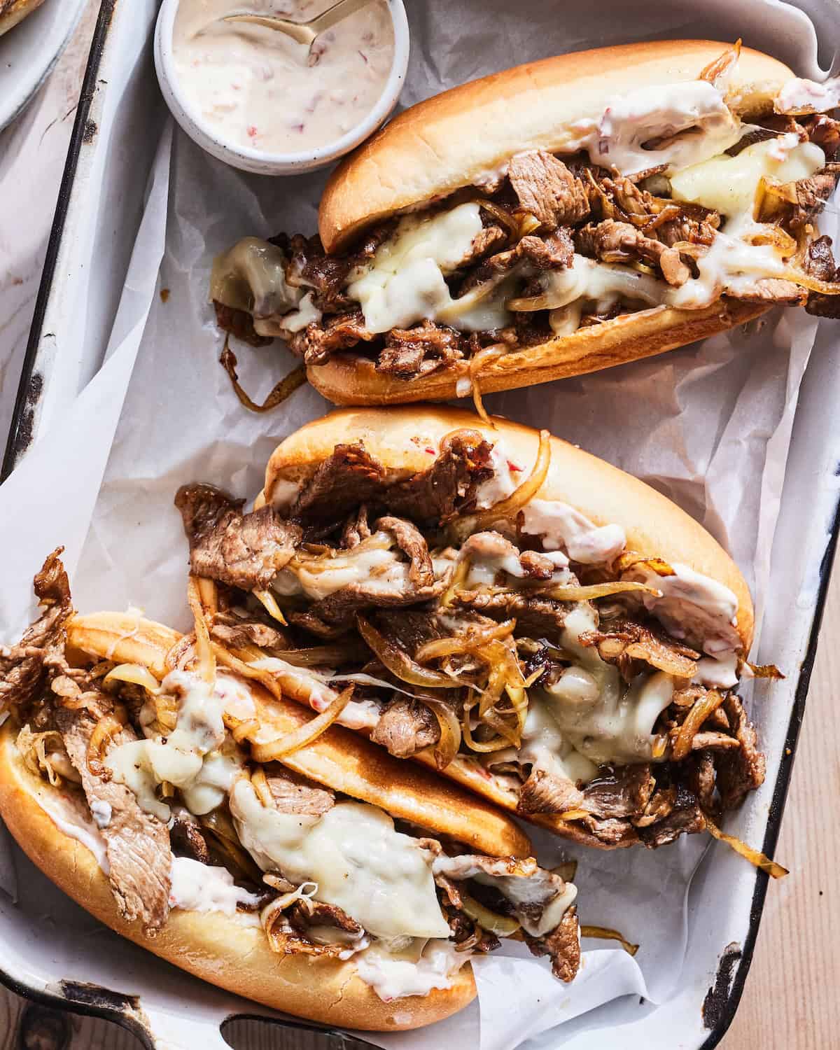 A close up shot of a rectangular dish with three philly cheesesteaks, along with a small bowl of cherry pepper mayonnaise.