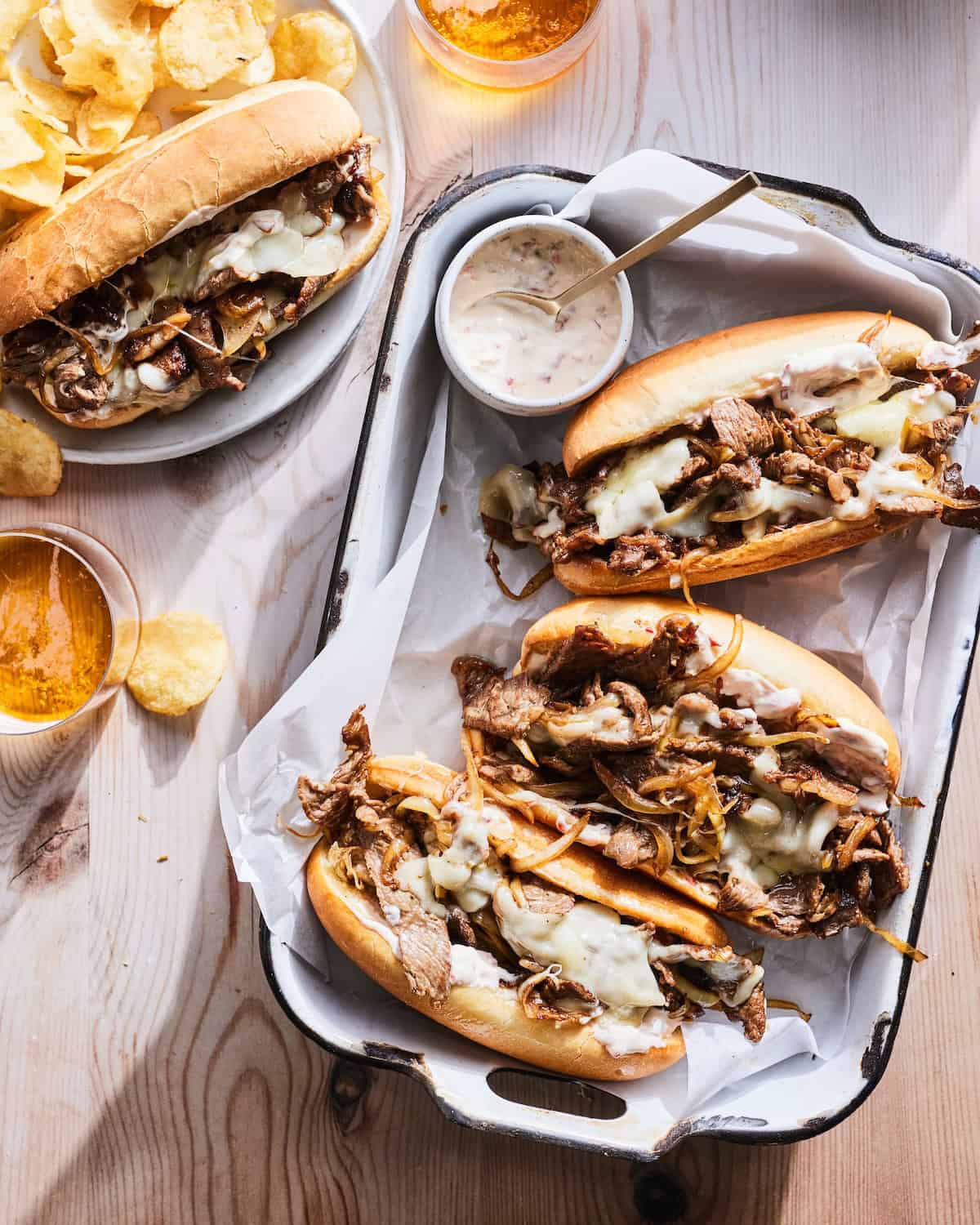 A rectangular dish with three philly cheesesteaks, along with a small bowl of cherry pepper mayonnaise, one cheesesteak served in a plate with potato chips and a couple beverages.