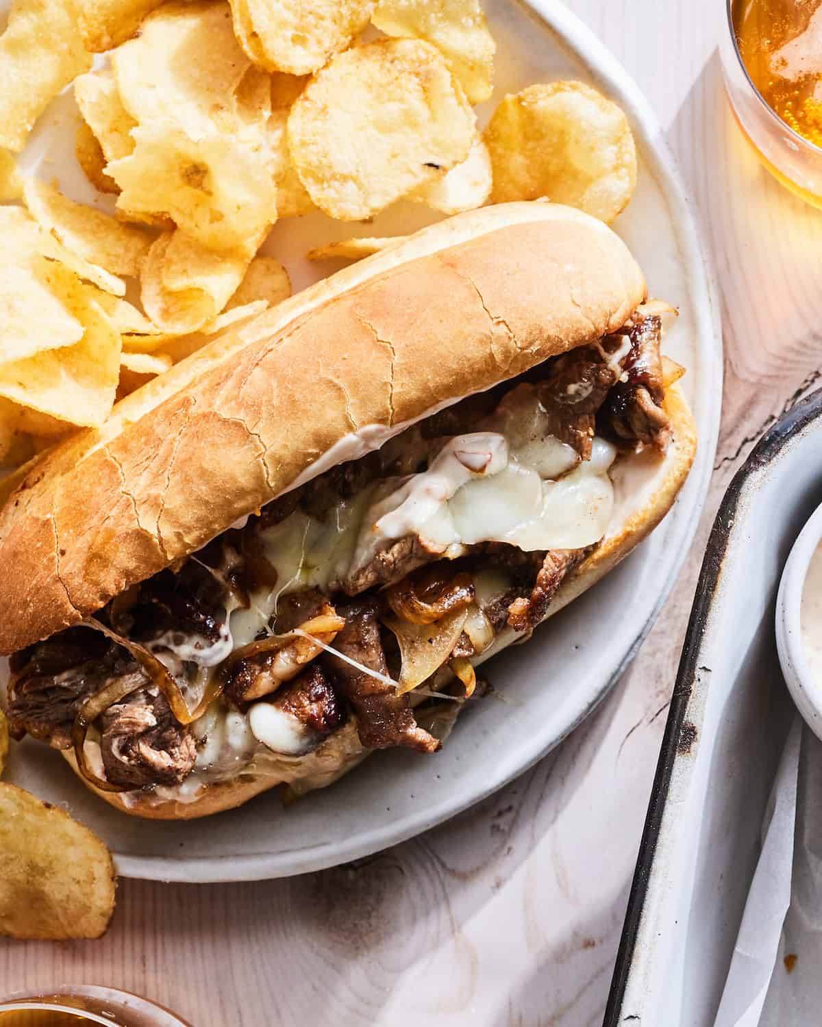 A close up shot of a philly cheesesteak served in a plate with potato chips and a beverage.