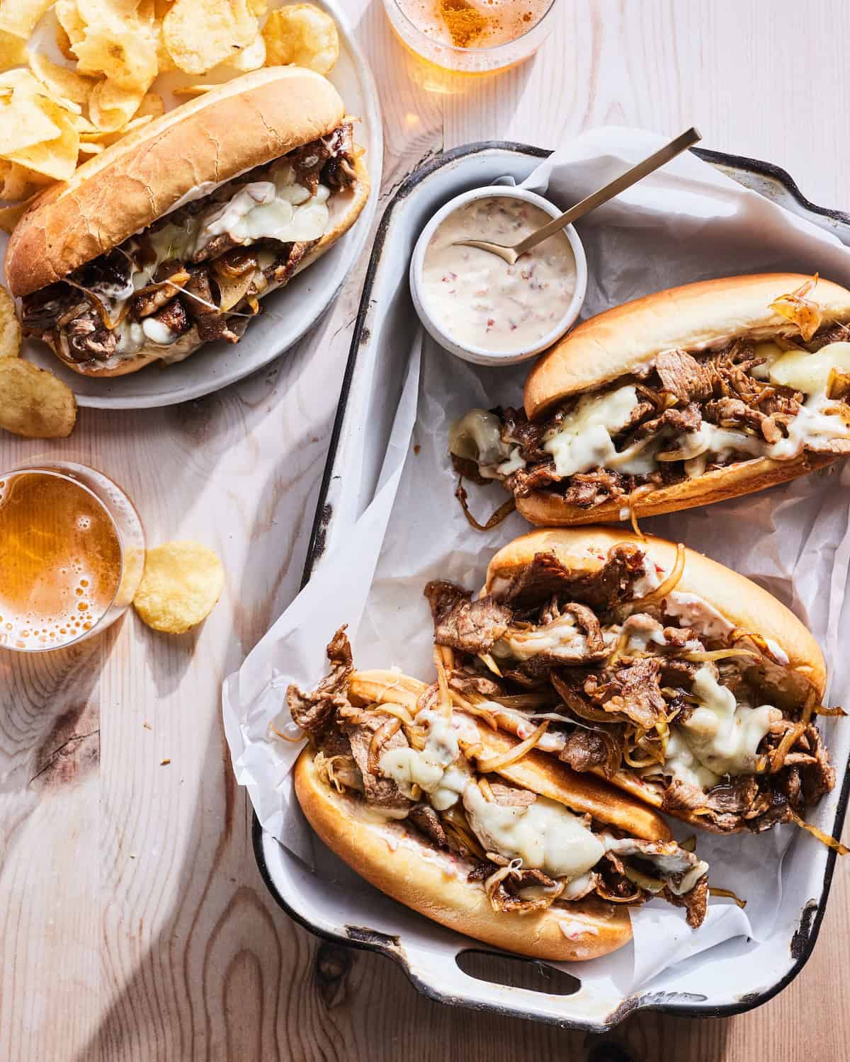 A rectangular dish with three philly cheesesteaks, along with a small bowl of cherry pepper mayonnaise, one cheesesteak served in a plate with potato chips and a couple beverages.