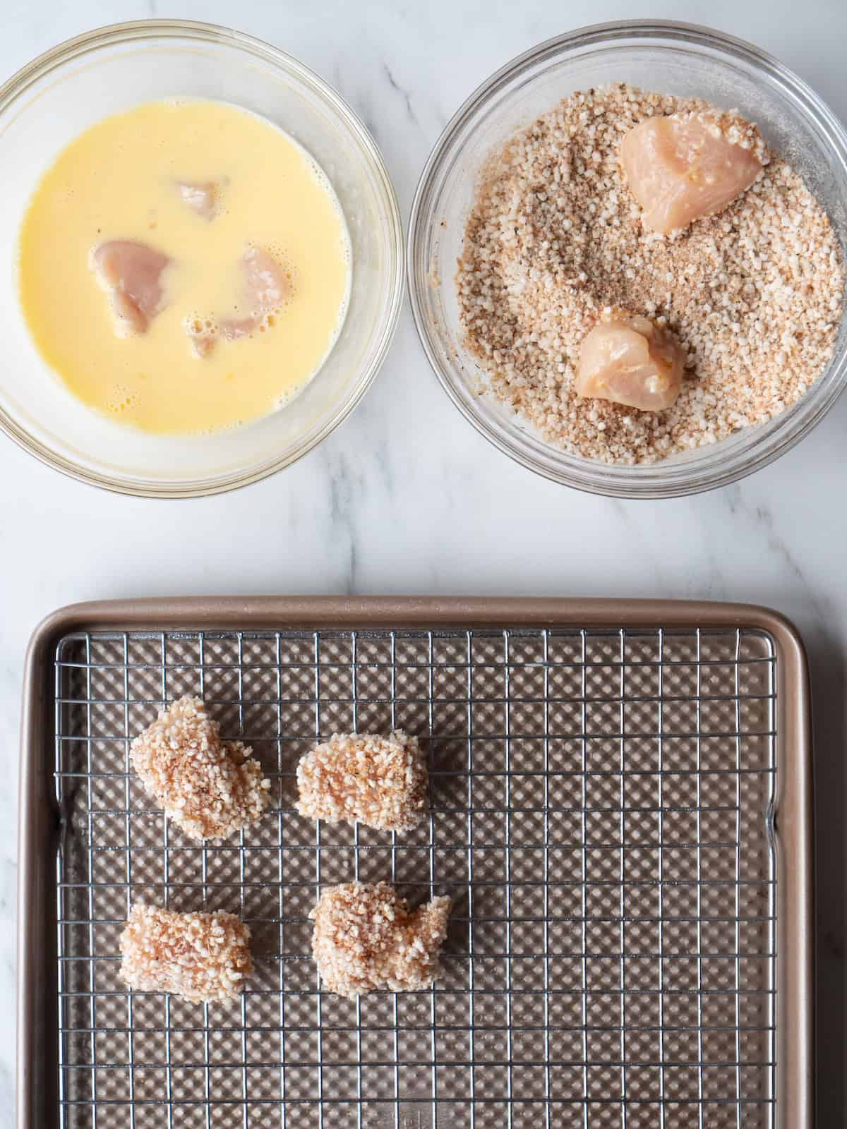A bowl with a milk and eggs mixture and another with seasoned panko breadcrumbs along with a baking sheet with a wire rack on it, with small pieces of chicken dipped in egg mix and coated in breadcrumbs placed on the wire rack.