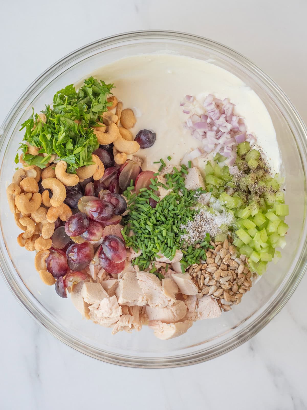 A mixing bowl with chicken salad dressing in the bottom topped with chicken, grapes, cashews, celery, parsley, chives, sunflower seeds, and shallots.