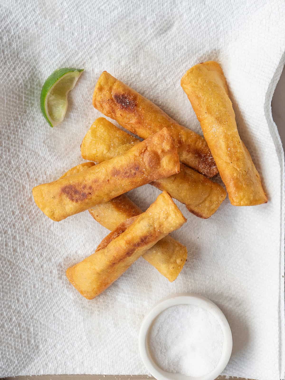 Fried Homemade Taquitos cooling on a paper towel lined plate with lime and sea salt