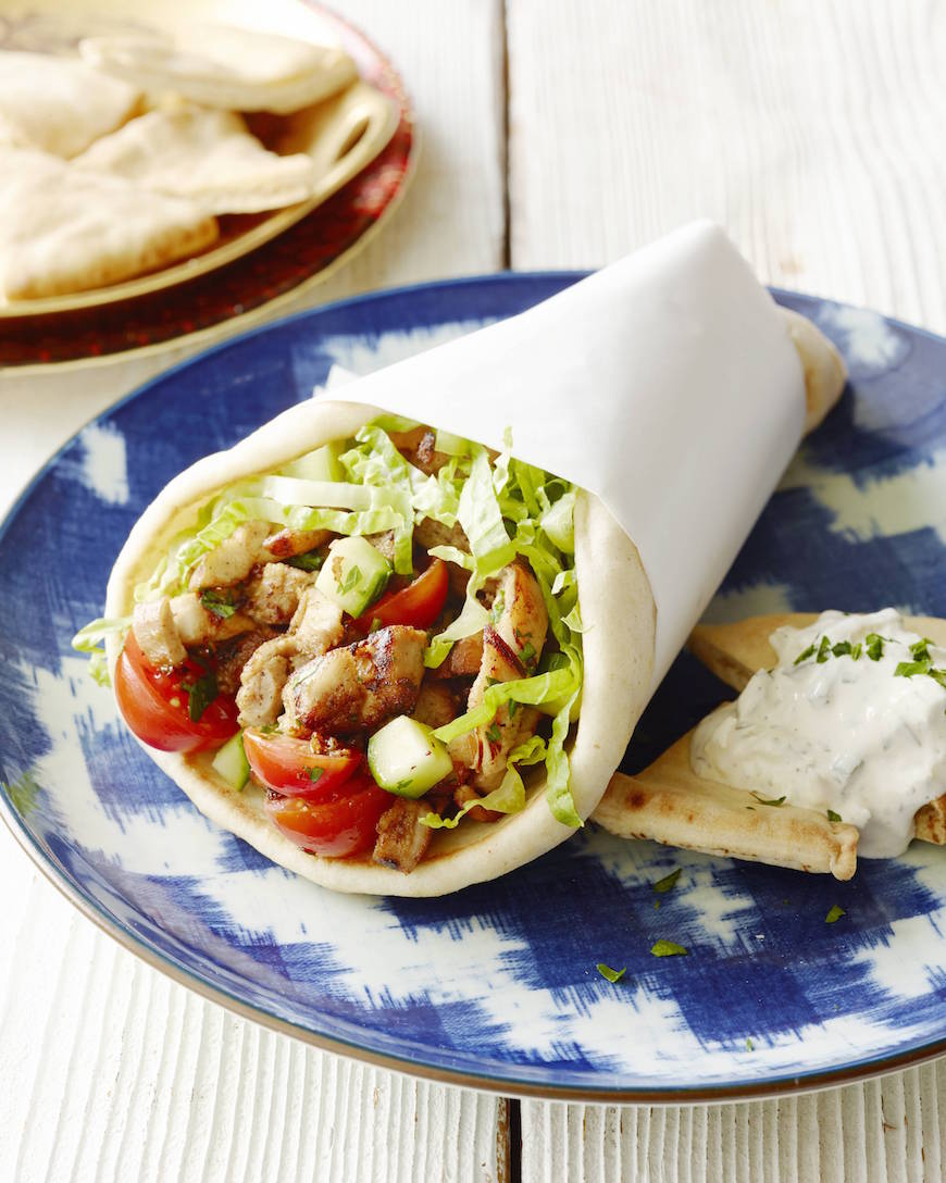 A chicken shawarma stuffed pita with a paper wrapped around to hold it, placed on a blue and white plate with tzatziki sauce and another plate with pita triangles in the background.